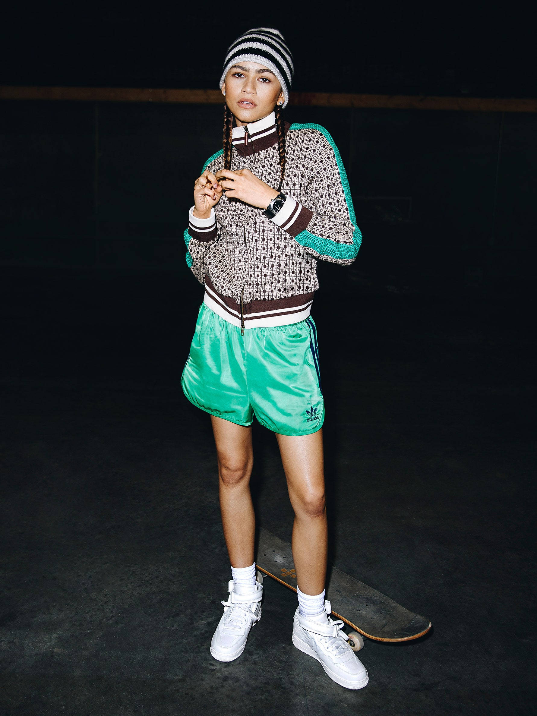 Zendaya 為 GQ 拍攝滑板少女造型 壁紙 — 一位時尚的年輕女性穿著圖案毛衣和亮綠色短褲，自信地站在一個光線昏暗的室內空間，腳邊放著滑板。
