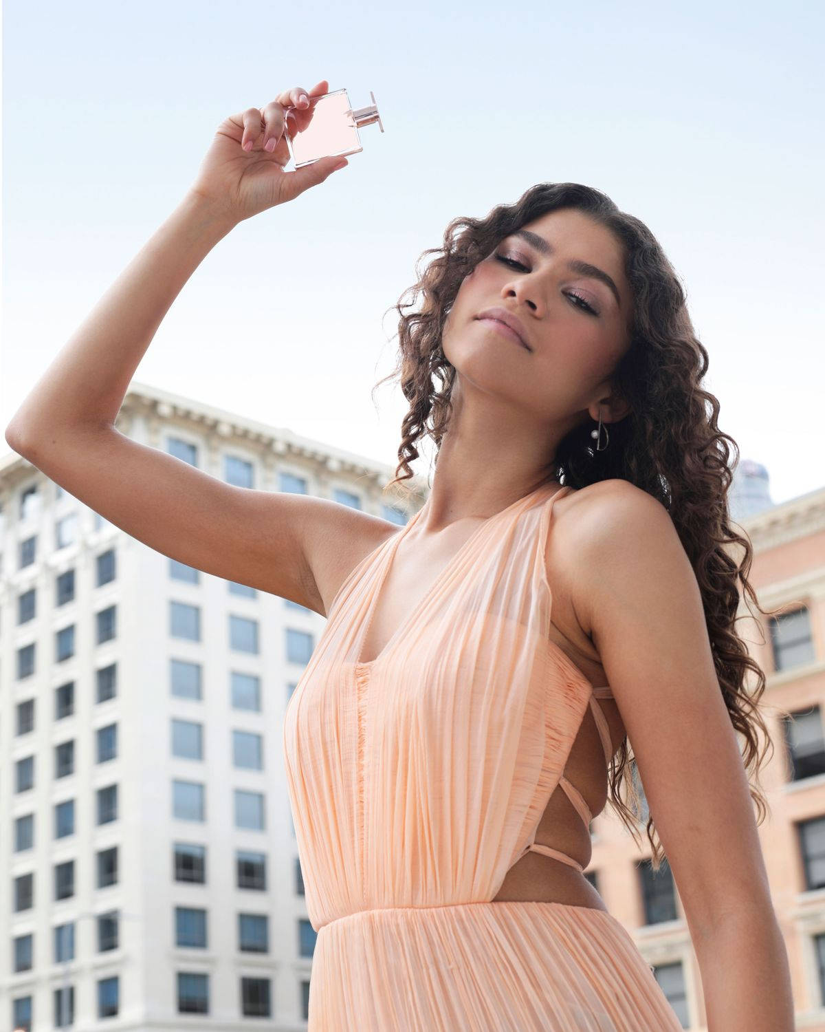 Zendaya 為 Lancôme Idole 拍攝香水廣告 壁紙 — 一位身穿桃色百褶裙的女性，在明亮的城市天際線前高舉香水瓶。