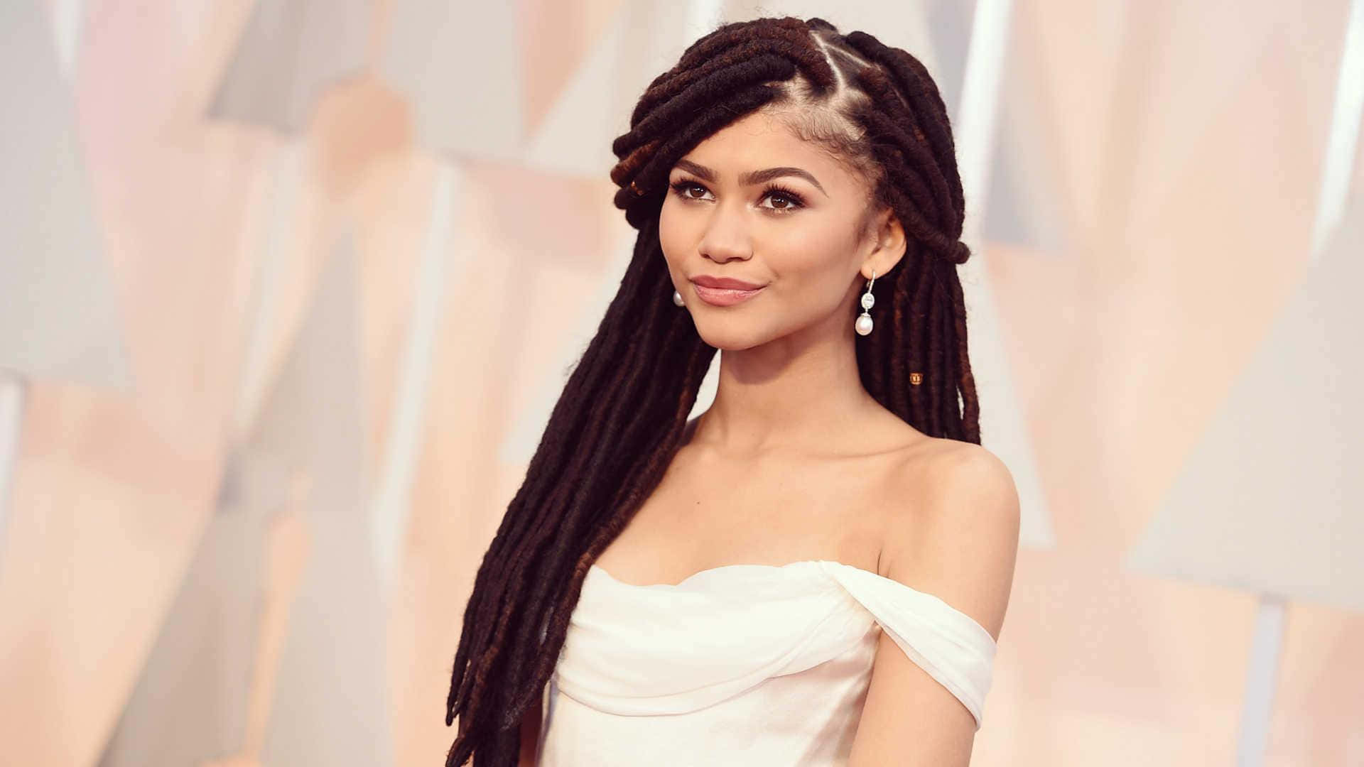 Zendaya 桌布 壁紙 — Zendaya 身穿白色露肩長裙，長辮子頭髮，在柔和的幾何背景前優雅地擺姿勢。