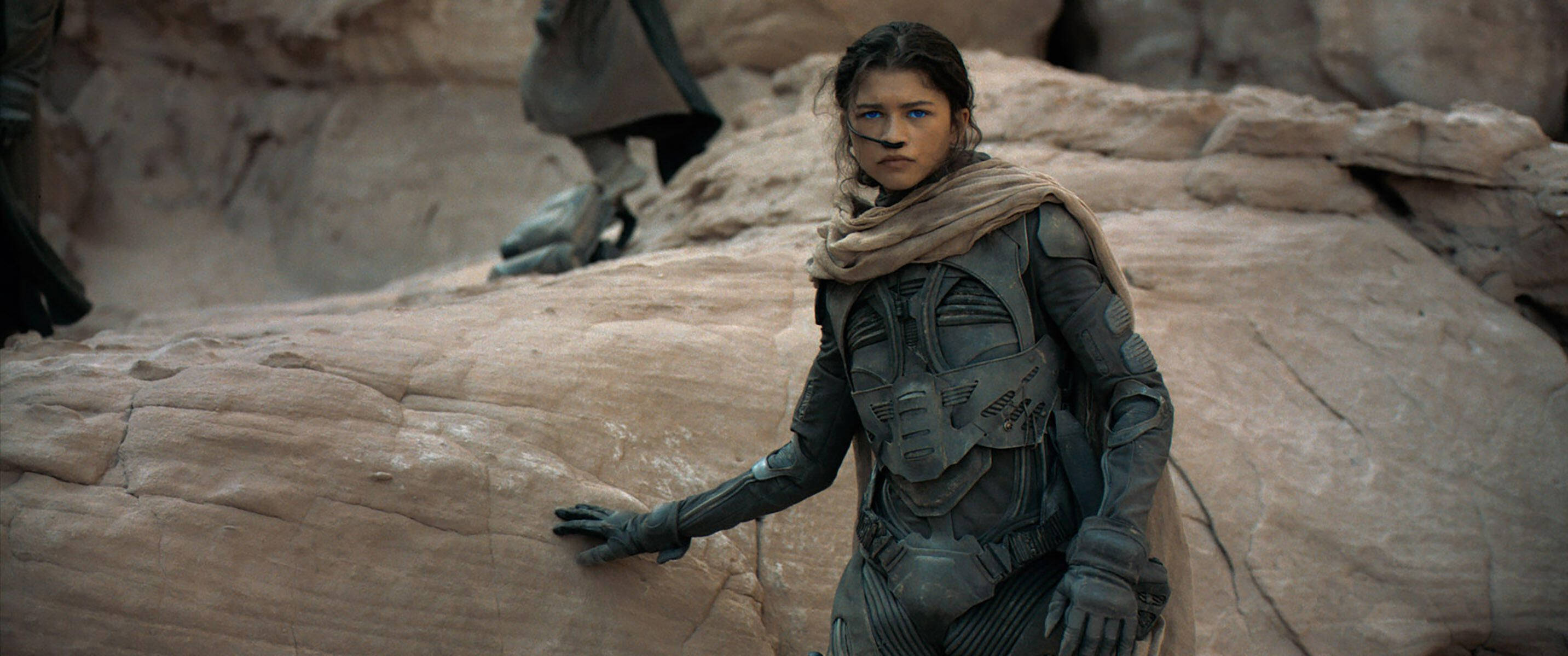 Zendaya 飾演欽尼 壁紙 — 一位堅定的女戰士身穿未來風格的盔甲，站在岩石地形上，身後是沙漠景觀。