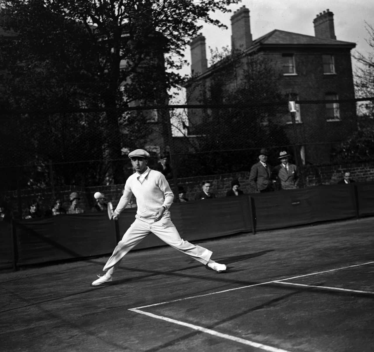法國商人雷內·拉科斯特 壁紙 — 一張復古黑白照片，一名網球運動員在戶外球場上全力揮拍，背景有觀眾和磚砌建築。