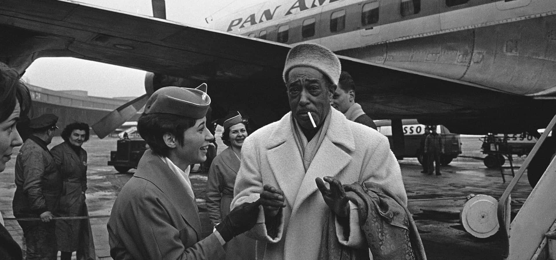  壁紙 — 一張復古黑白照片顯示，一位微笑的泛美航空空服員在機場停機坪上，向一位穿著大衣、戴著帽子、抽著香煙的男子打招呼。