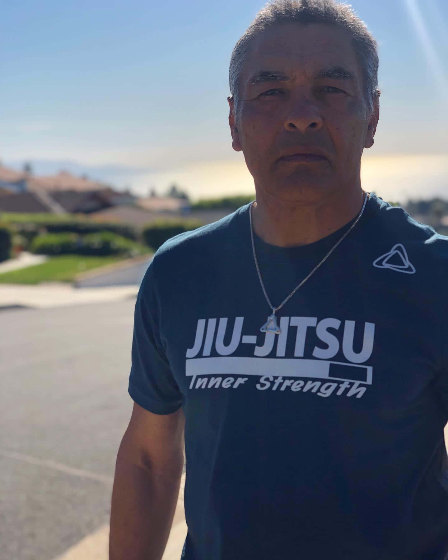  壁紙 — 一位中年男子穿著深藍色柔術T恤，在明亮的陽光下戶外站立，背景是郊區住宅區。