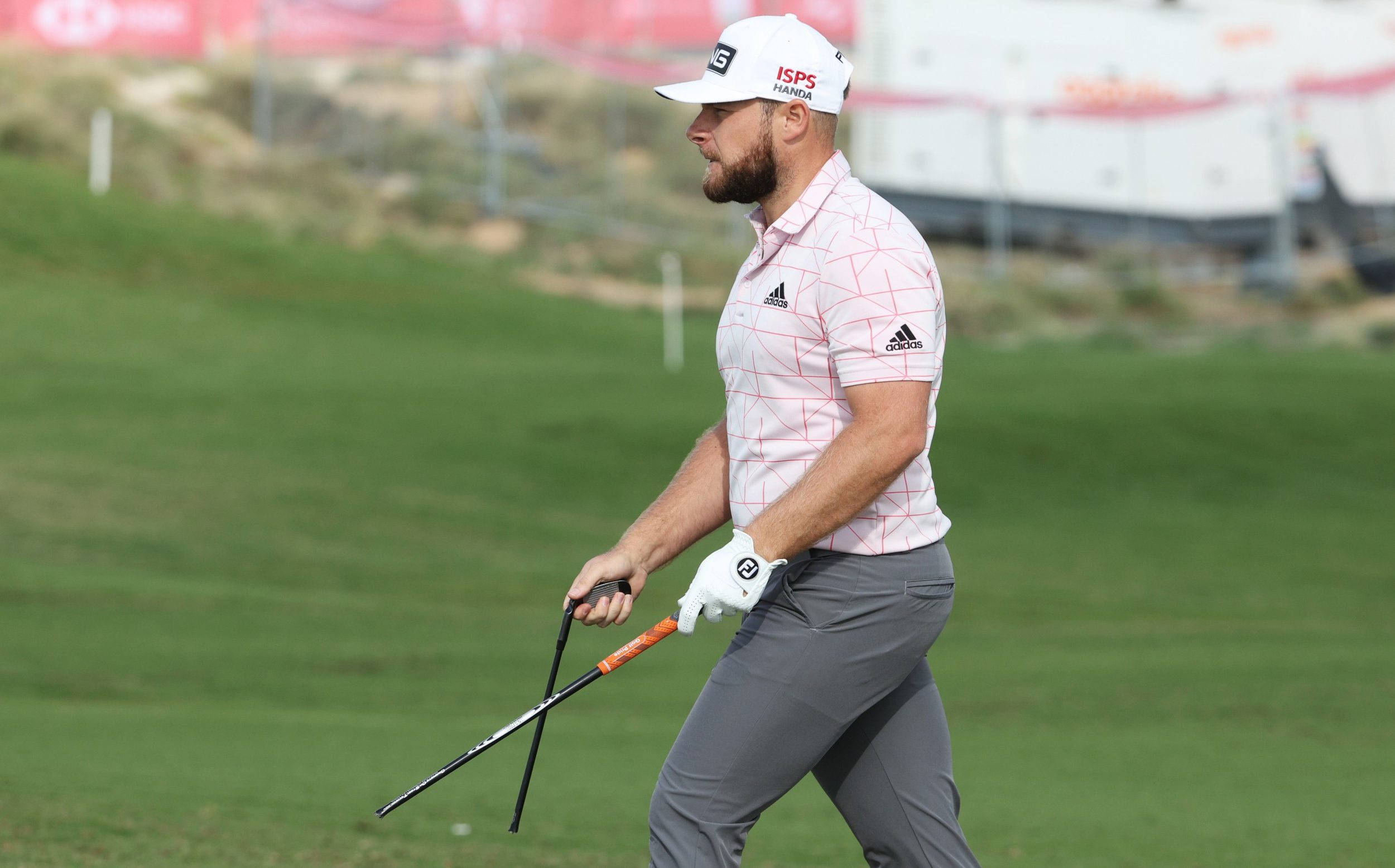 Tyrrell Hatton 桌布 壁紙 — 一位專注的高爾夫球手穿著粉紅色圖案襯衫和白色帽子，在翠綠的球道上拿著高爾夫球桿行走。