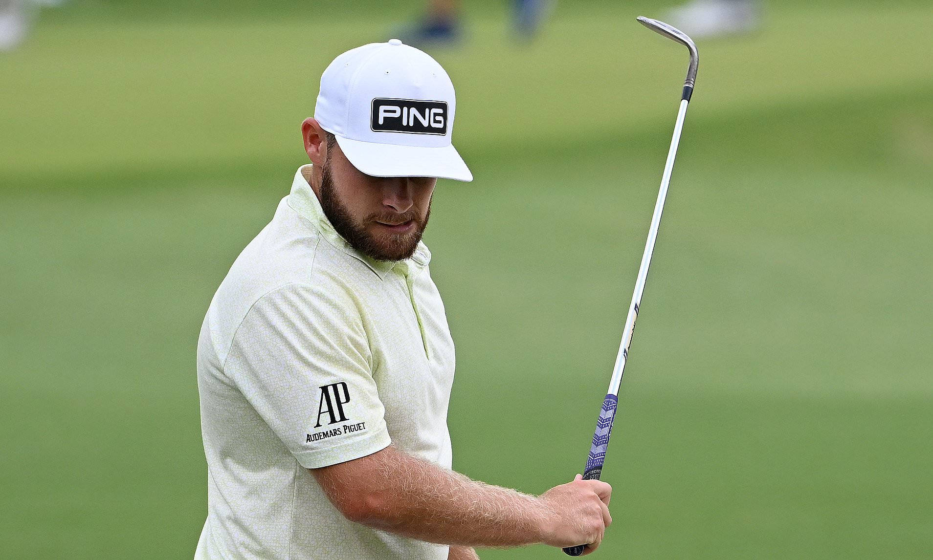 Tyrrell Hatton 桌布 壁紙 — 一位專注的高爾夫球手，戴著白色帽子，穿著Polo衫，在翠綠的球場上握著球桿。