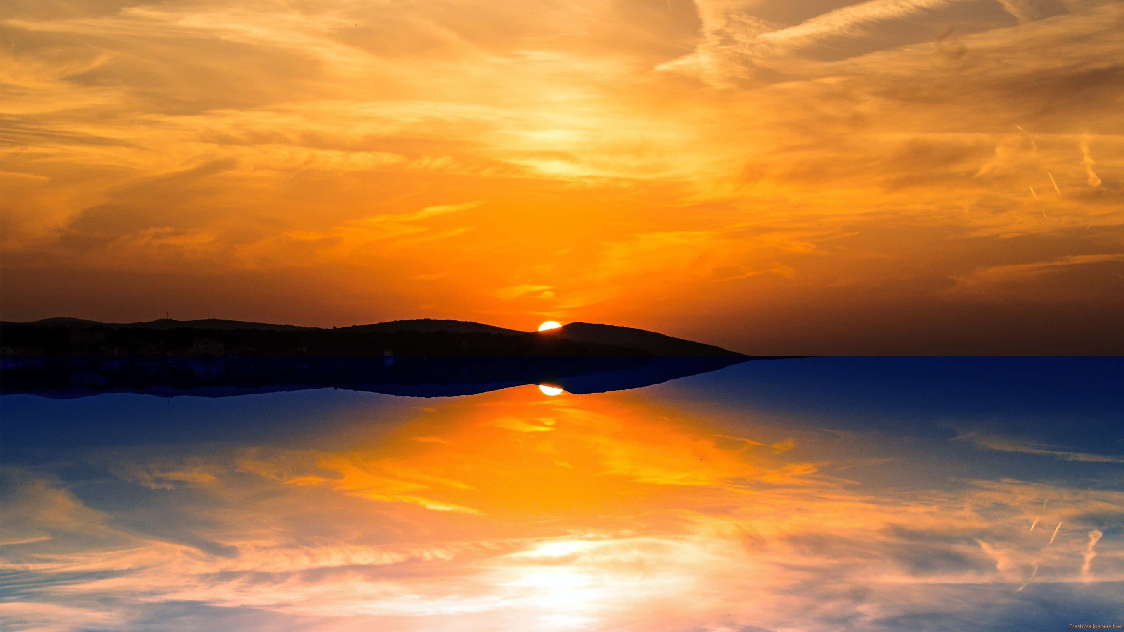 Sunset Reflection 桌布 壁紙 — 壯麗的日落，火橙色的雲彩完美地倒映在平靜的水面上，下方是剪影般的山丘。