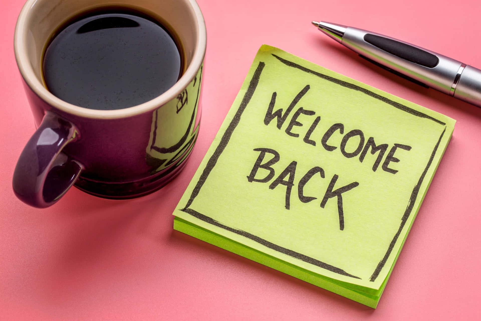  壁紙 — 一個紫色咖啡杯旁邊放著一張寫有「WELCOME BACK」的亮綠色便利貼，還有一支銀色筆在粉紅色表面上。