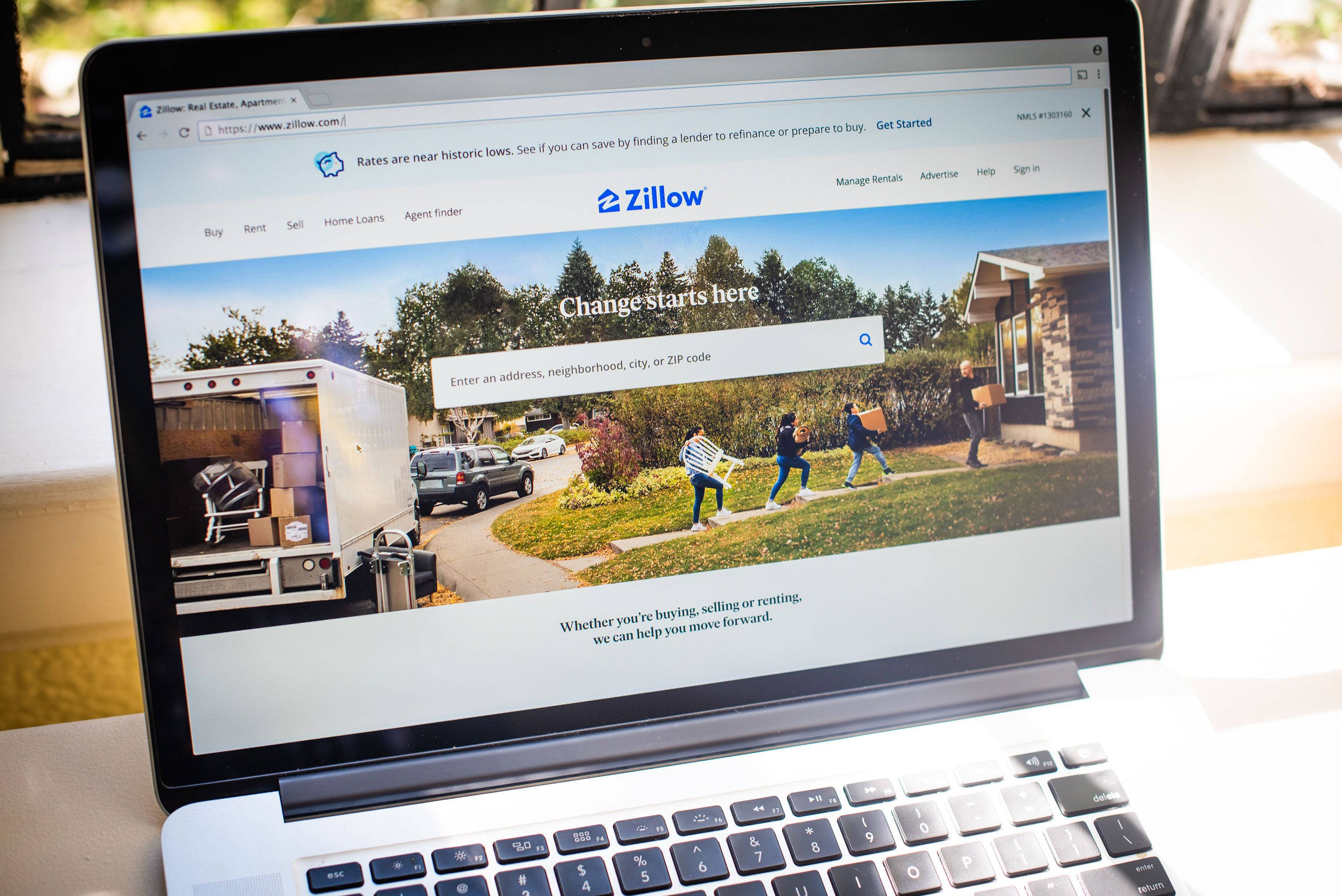 筆記本電腦顯示 Zillow 房地產網站 壁紙 — 手提電腦顯示Zillow房地產首頁，明亮的郊區背景下有移動的人和卡車。