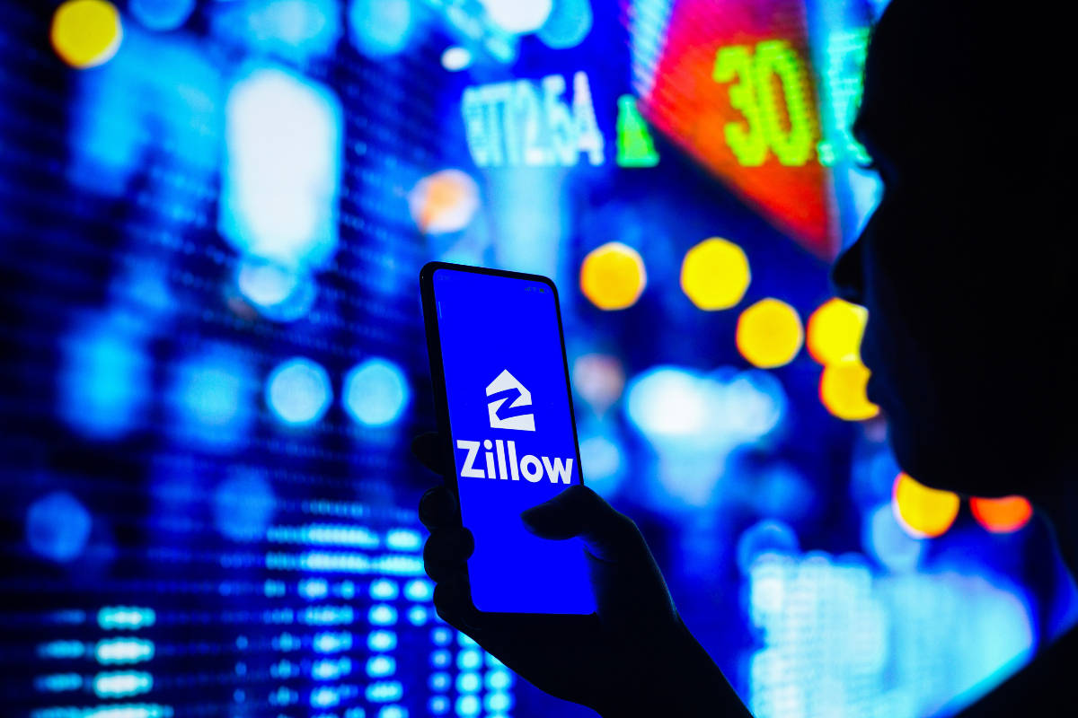 Zillow與彩色城市燈光 壁紙 — 一個人嘅剪影，揸住部顯示Zillow應用程式嘅電話，背景係充滿活力嘅模糊股票市場螢幕。