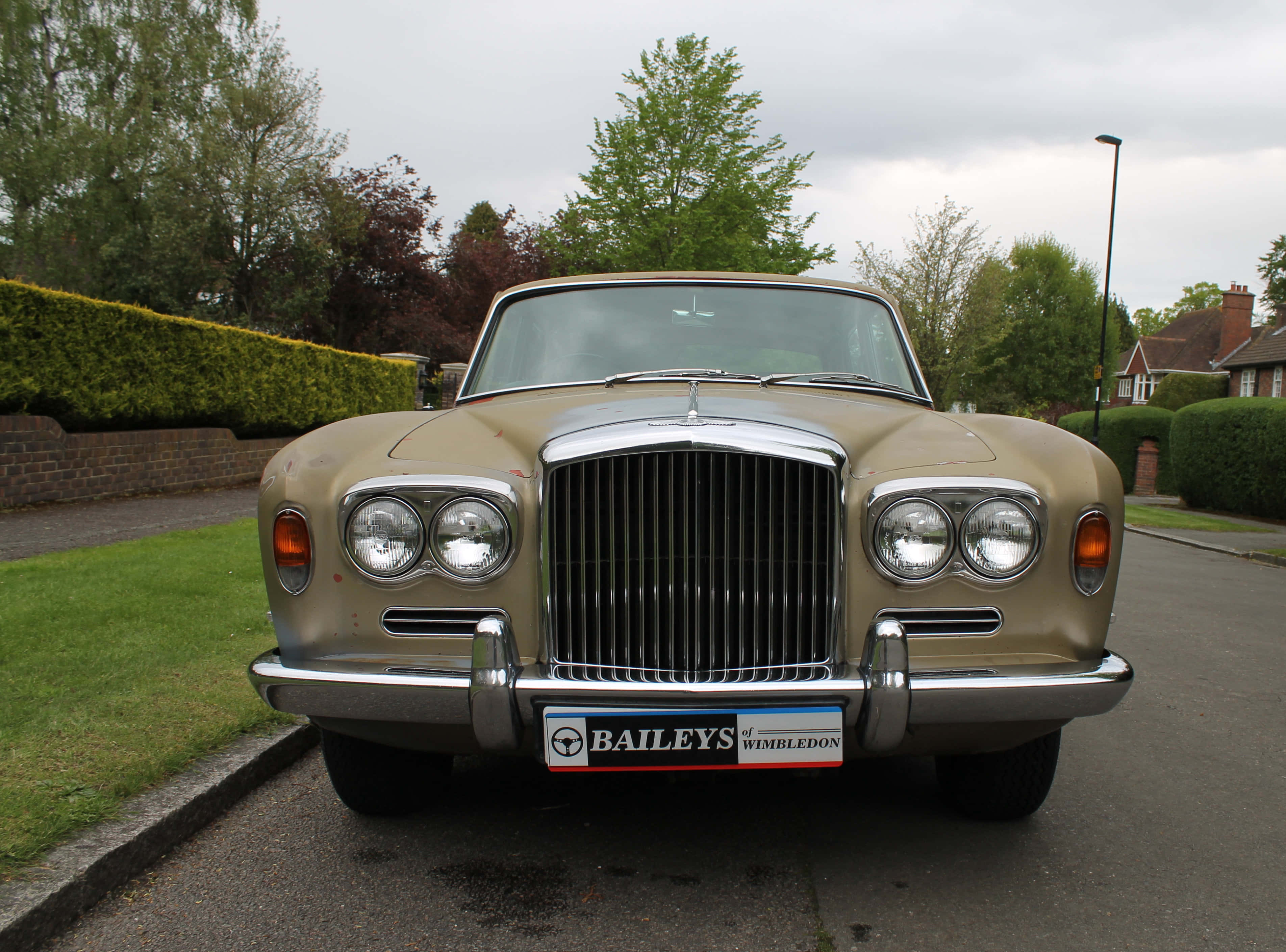 Bentley T1 桌布 壁紙 — 一輛棕褐色復古賓利，帶有鍍鉻格柵和圓形頭燈，停泊在郊區街道上，天空多雲。