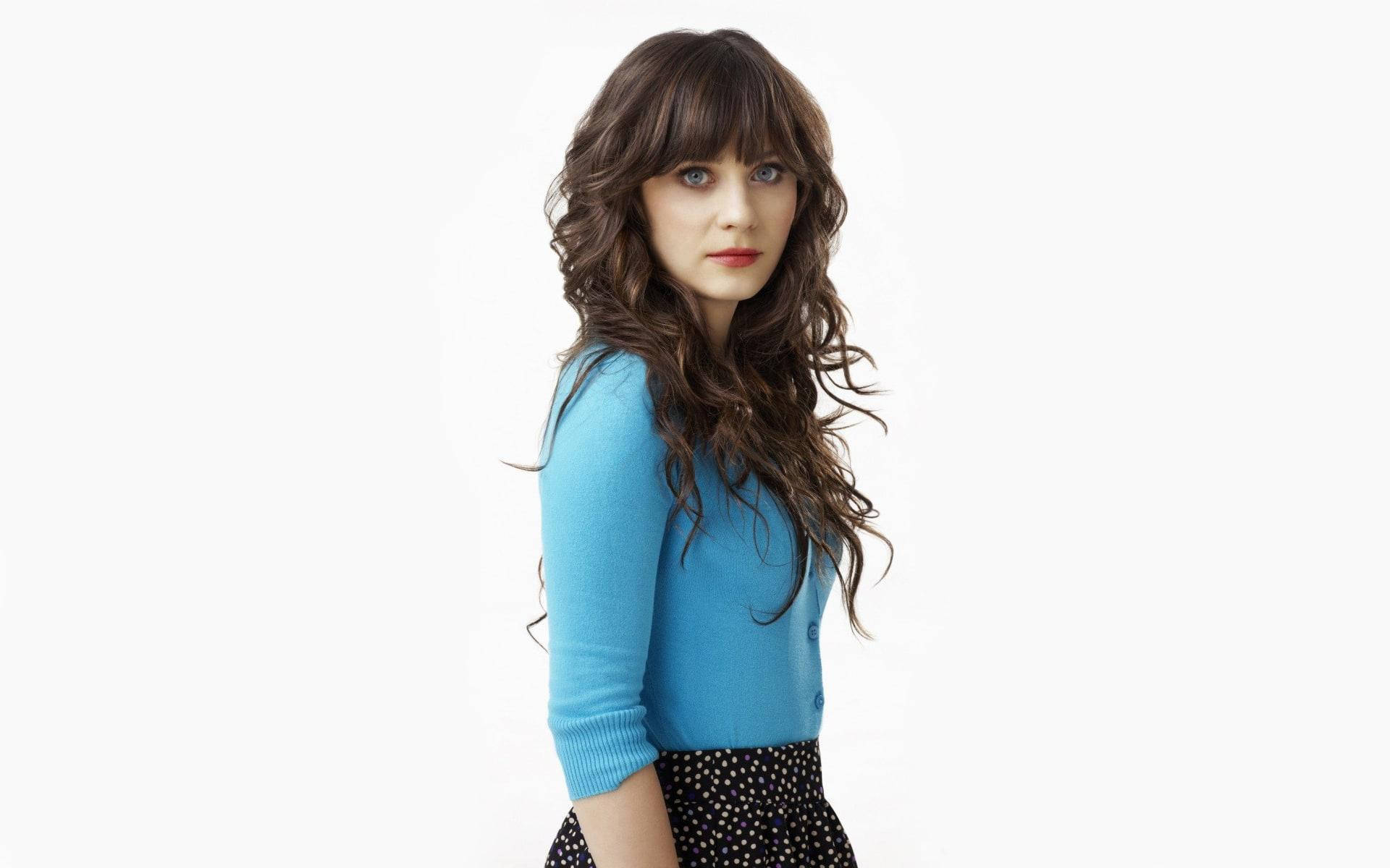Zooey Deschanel 穿藍色學院風服裝 壁紙 — 一位擁有波浪棕色頭髮和瀏海的女性，穿著藍色毛衣和圓點裙，背景是純白色。