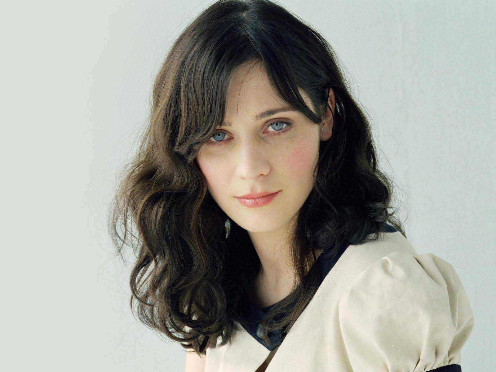 Zooey Deschanel 白色美學寫真 壁紙 — 一位擁有波浪深色頭髮和引人注目的藍眼睛的女性，在純淺灰色背景下柔和地凝視鏡頭。