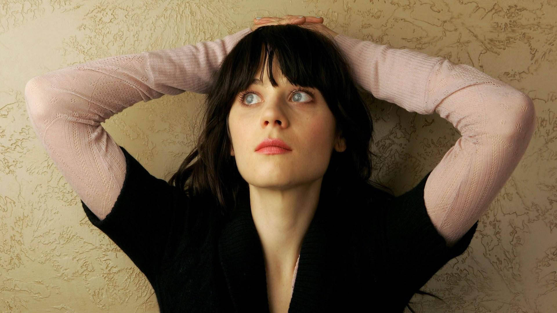 Zooey Deschanel 穿黑粉毛衣 壁紙 — 一位深色頭髮、擁有引人注目的藍眼睛的女性，雙手交握在腦後，靠著紋理米色牆壁向上看。