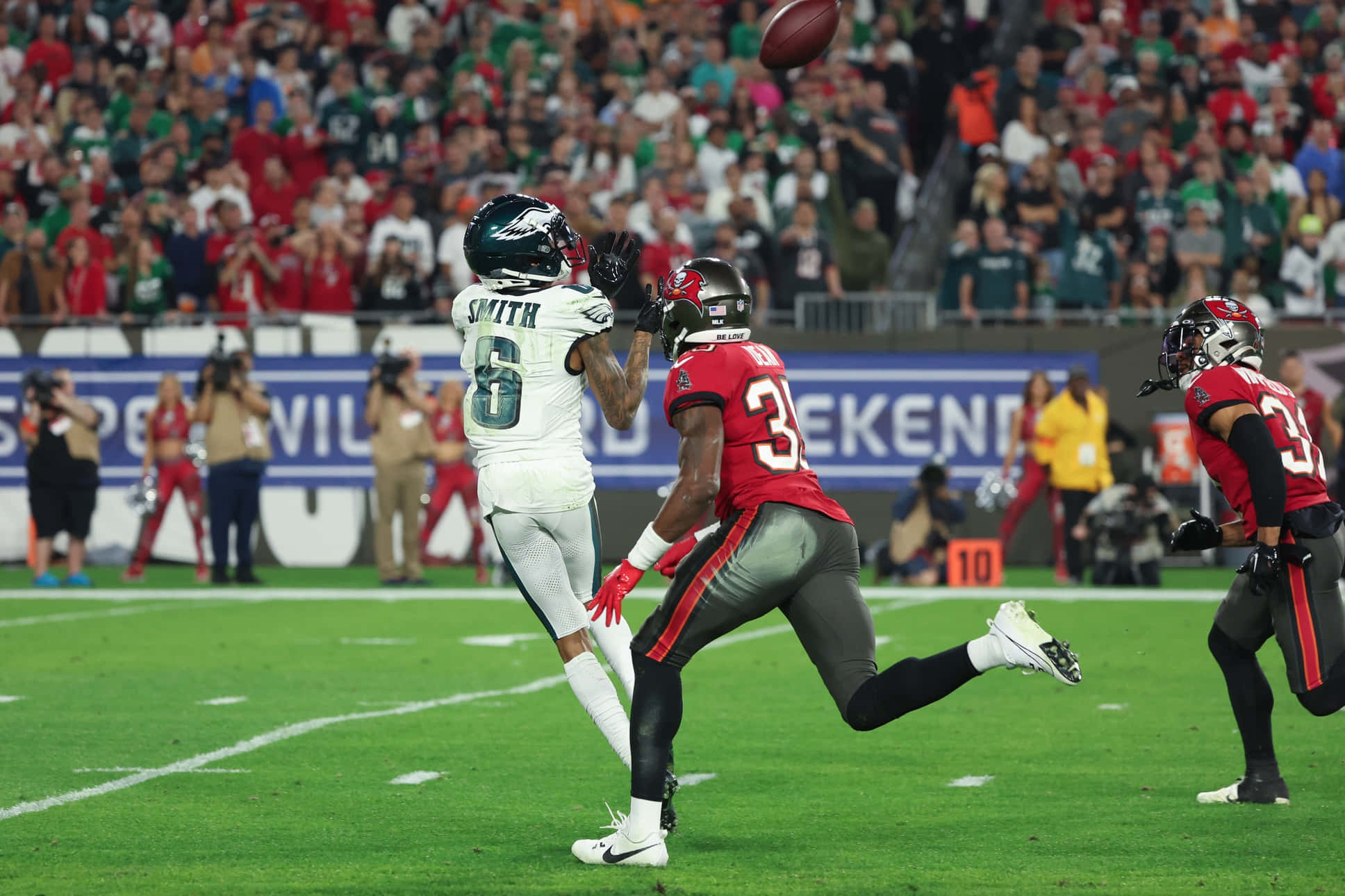 Devonta Smith 桌布 壁紙 — 一名身穿白色球衣的足球員跳起接球，而一名身穿紅色球衣的防守球員在座無虛席的體育館比賽中逼近。