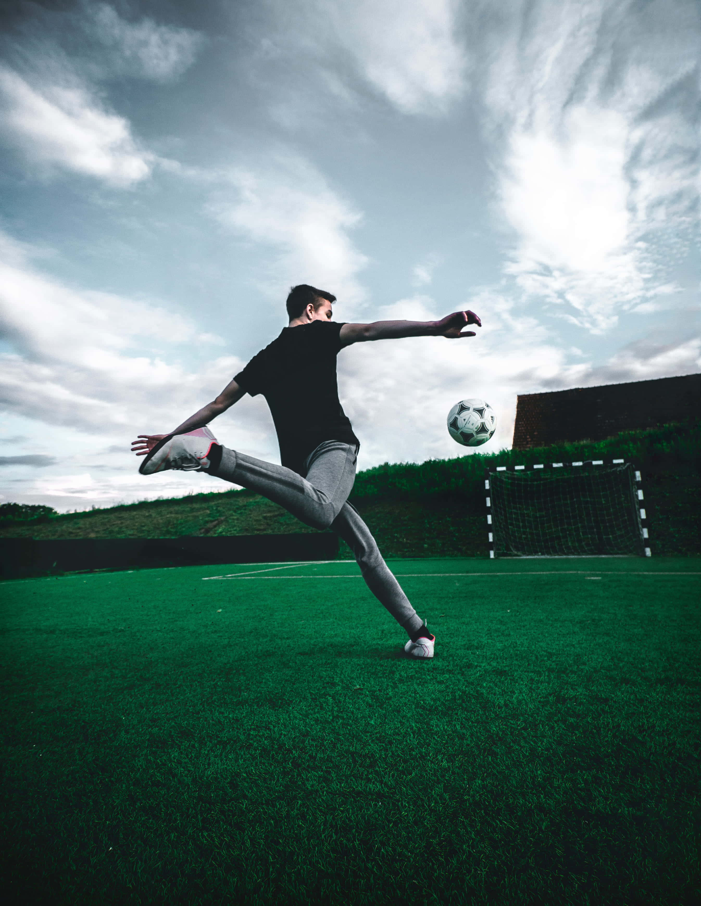 Best Sports 桌布 壁紙 — 一名足球運動員在充滿活力的綠色球場上踢球，背景是戲劇性的多雲天空，遠處有一個球門。