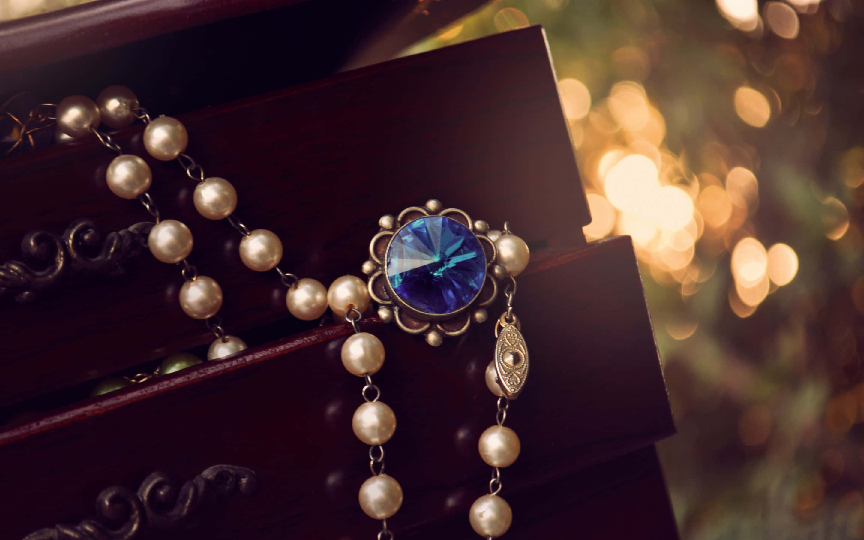 Jewelry Design 桌布 壁紙 — 一串帶有藍色水晶中心裝飾的珍珠玫瑰經，優雅地放置在深色木質表面上，背景是溫暖的散景效果。