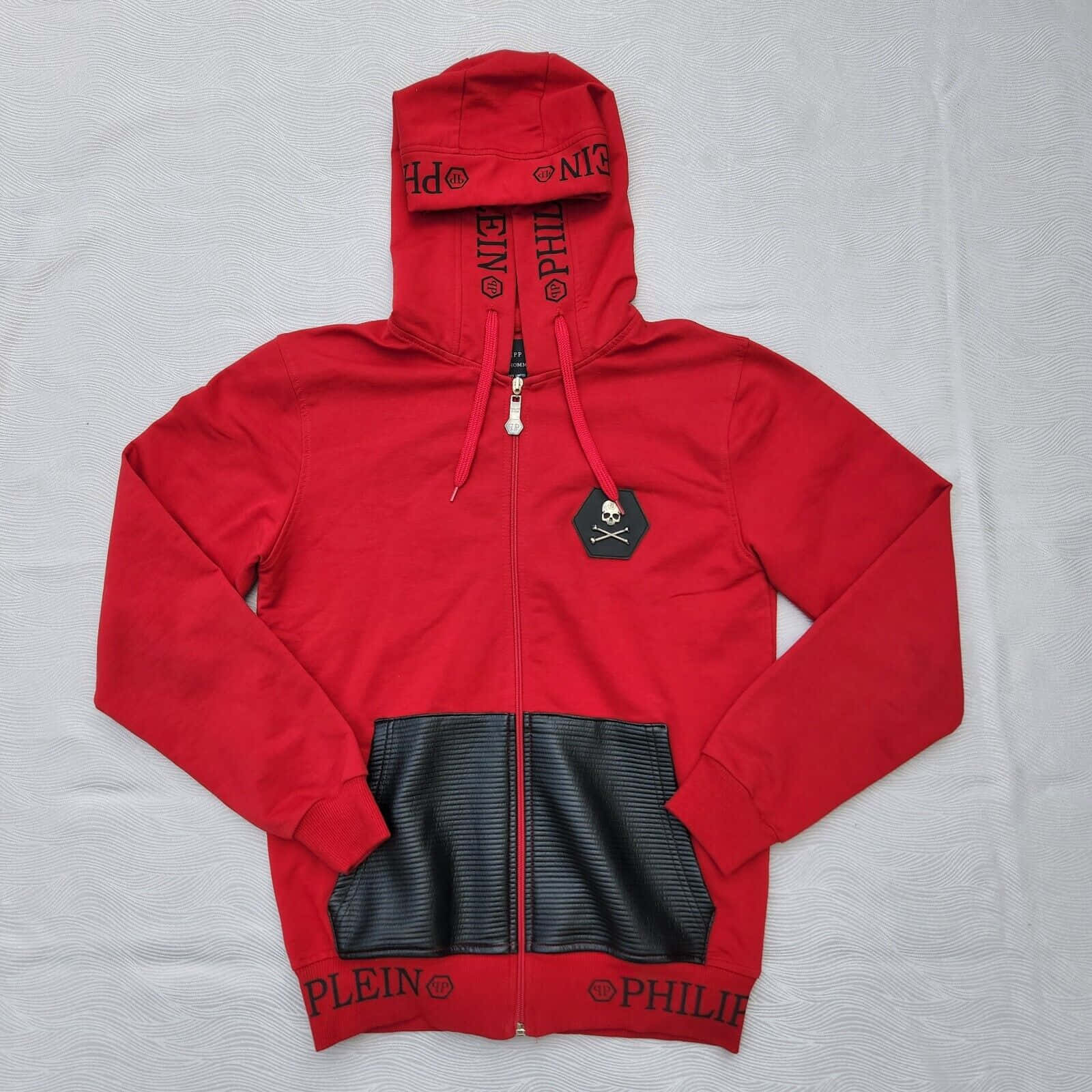 Red Hoodie 桌布 壁紙 — 一件大膽的紅色 Philipp Plein 拉鏈連帽衛衣，帶有黑色皮革拼接和骷髏標誌，平放在有紋理的白色表面上。