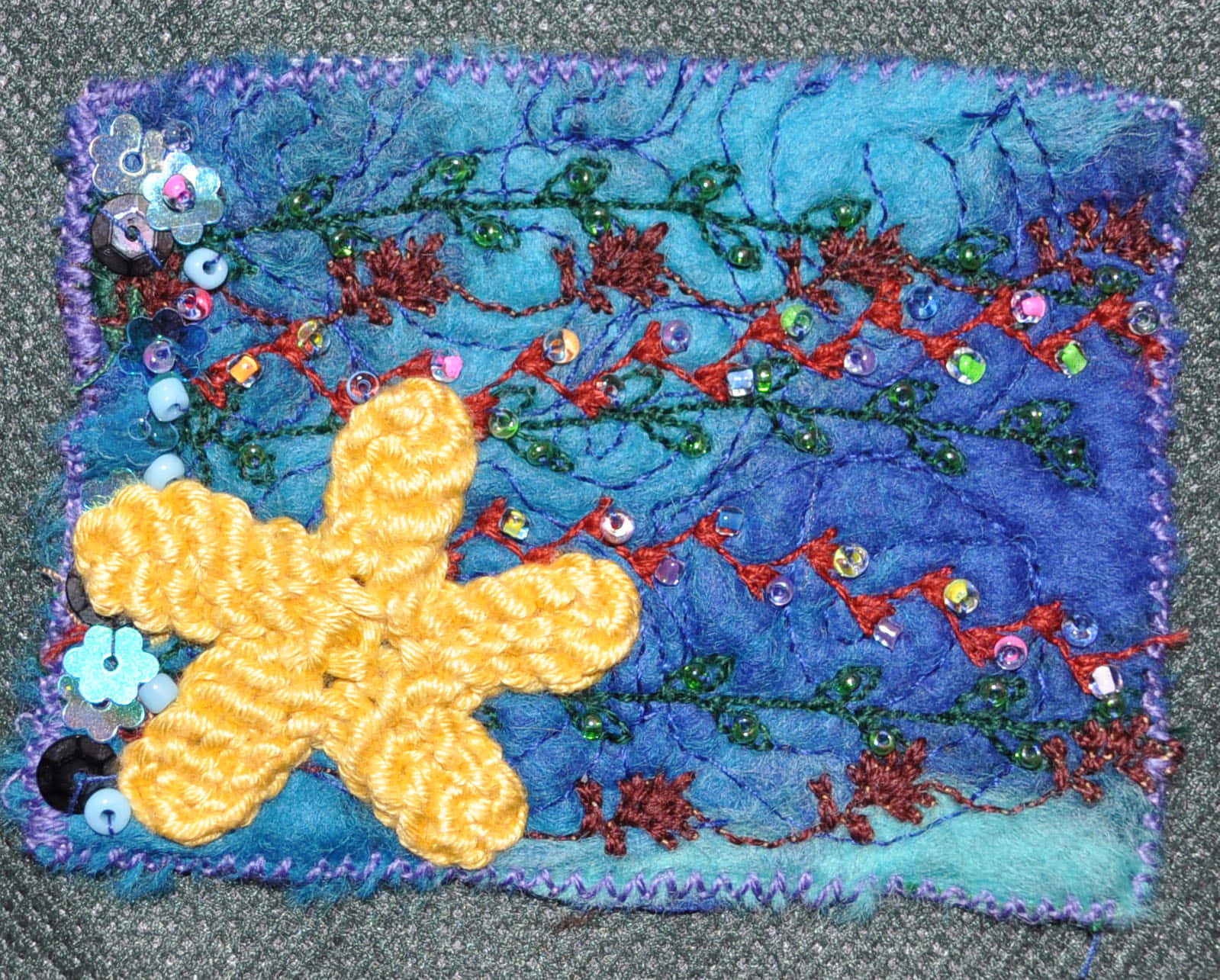Textile Art 桌布 壁紙 — 一件手工紡織藝術品，在藍色紋理布料上有一個黃色鉤針編織的海星，飾有紅綠色線藤蔓和彩色珠子。