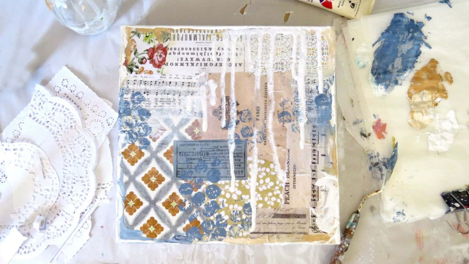 Mixed Media 桌布 壁紙 — 一件混合媒介拼貼藝術作品，帶有花卉圖案、復古文字和顏料滴落，放置在一塊白布上，周圍環繞著蕾絲墊和畫筆。