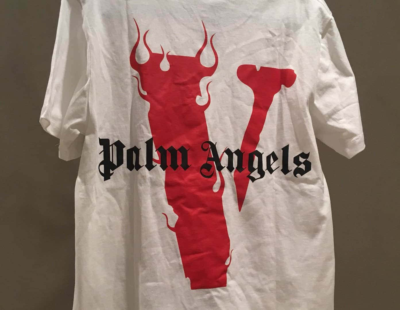 Vlone 桌布 壁紙 — 一件白色T恤，帶有醒目的紅色惡魔角 'V' 標誌和黑色 'Palm Angels' 文字，掛在純棕色牆壁上。