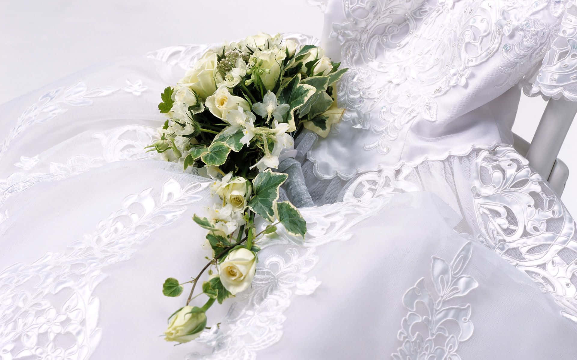 Wedding Flowers 桌布 壁紙 — 一件精緻的白色婚紗，帶有複雜的蕾絲細節，在柔和的白色背景上托著一束奶油色玫瑰和雜色常春藤的瀑布式花束。