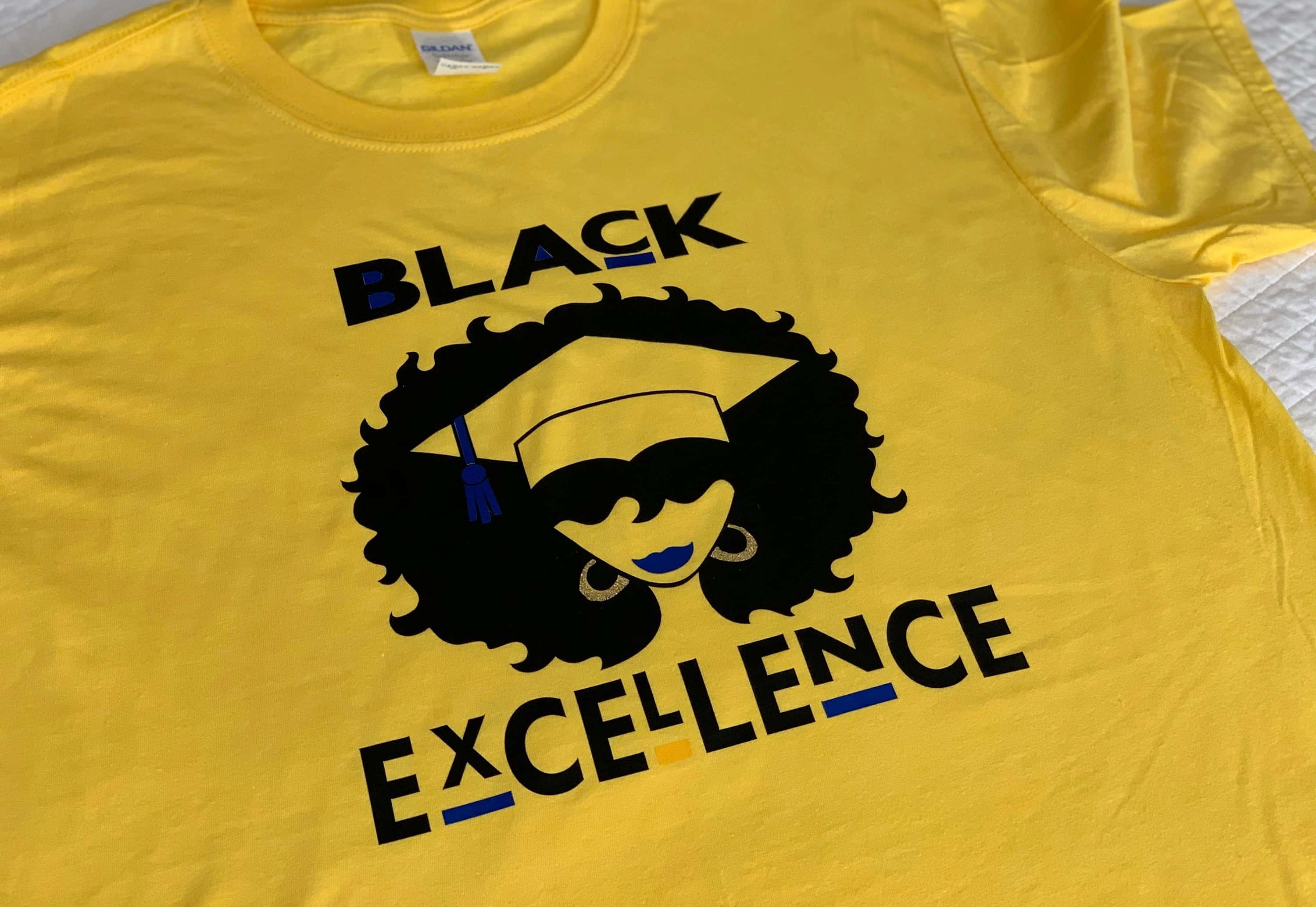Black Excellence 桌布 壁紙 — 一件鮮黃色T恤上印有醒目黑色圖案，圖案為一位面帶微笑、留著非洲式髮型、戴著畢業帽的女性，並有「BLACK EXCELLENCE」字樣。