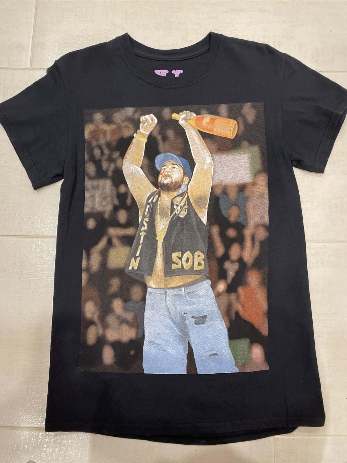 Vlone 桌布 壁紙 — 一件黑色T恤，上面有鮮豔的圖案，描繪一個戴藍色帽子和背心的男人在音樂會上高舉瓶子，平放在瓷磚地板上。