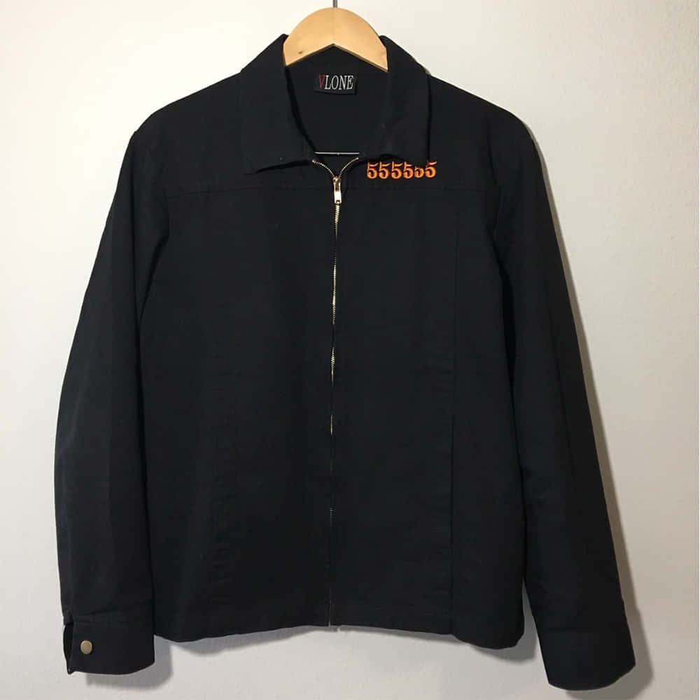 Vlone 桌布 壁紙 — 一件黑色Vlone外套帶有橙色泰文，整齊地掛在木衣架上，背景是素牆。