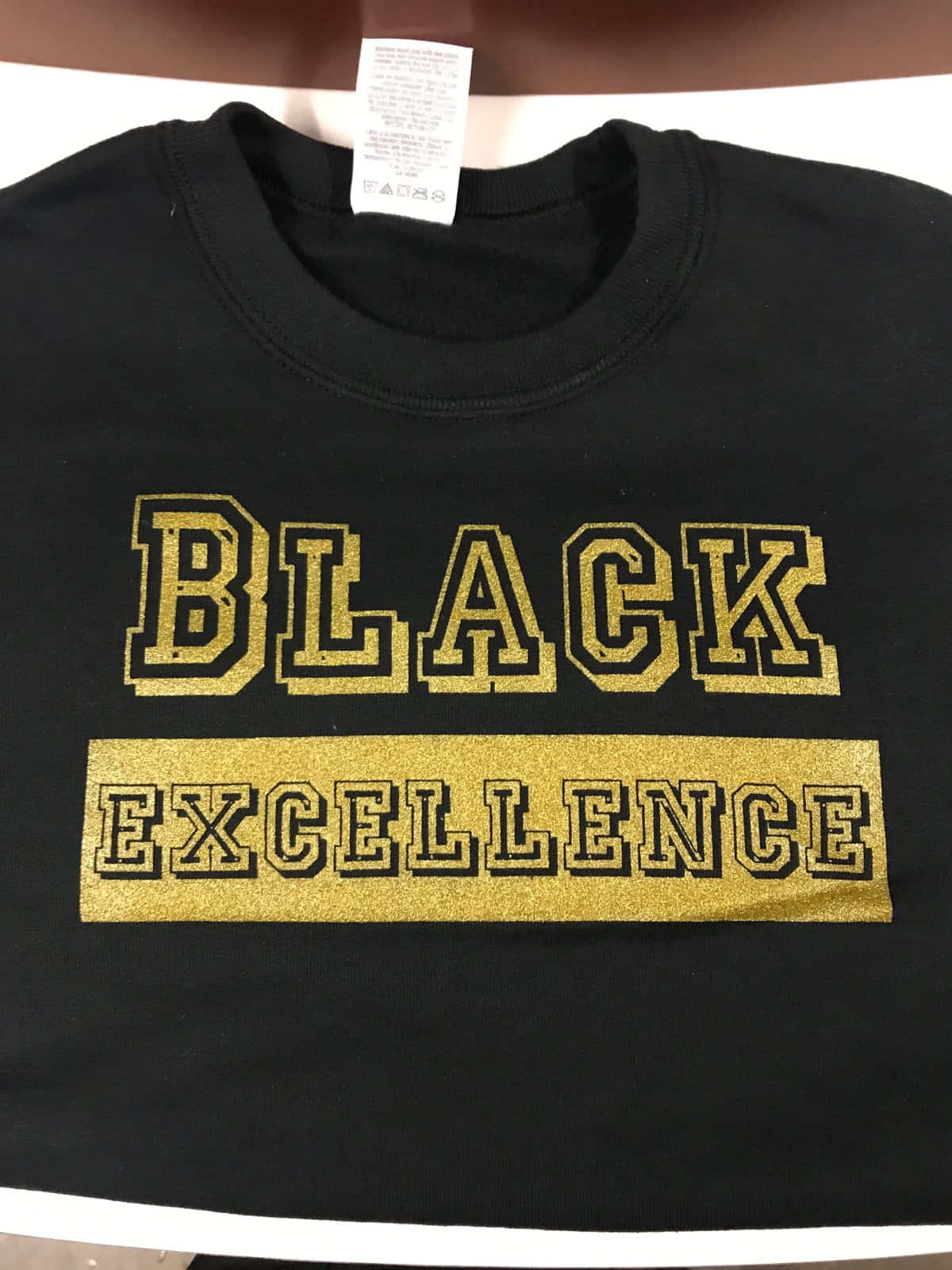 Black Excellence 桌布 壁紙 — 一件黑色衛衣，上面有粗體金色閃粉文字寫著 BLACK EXCELLENCE，展示在平面上。