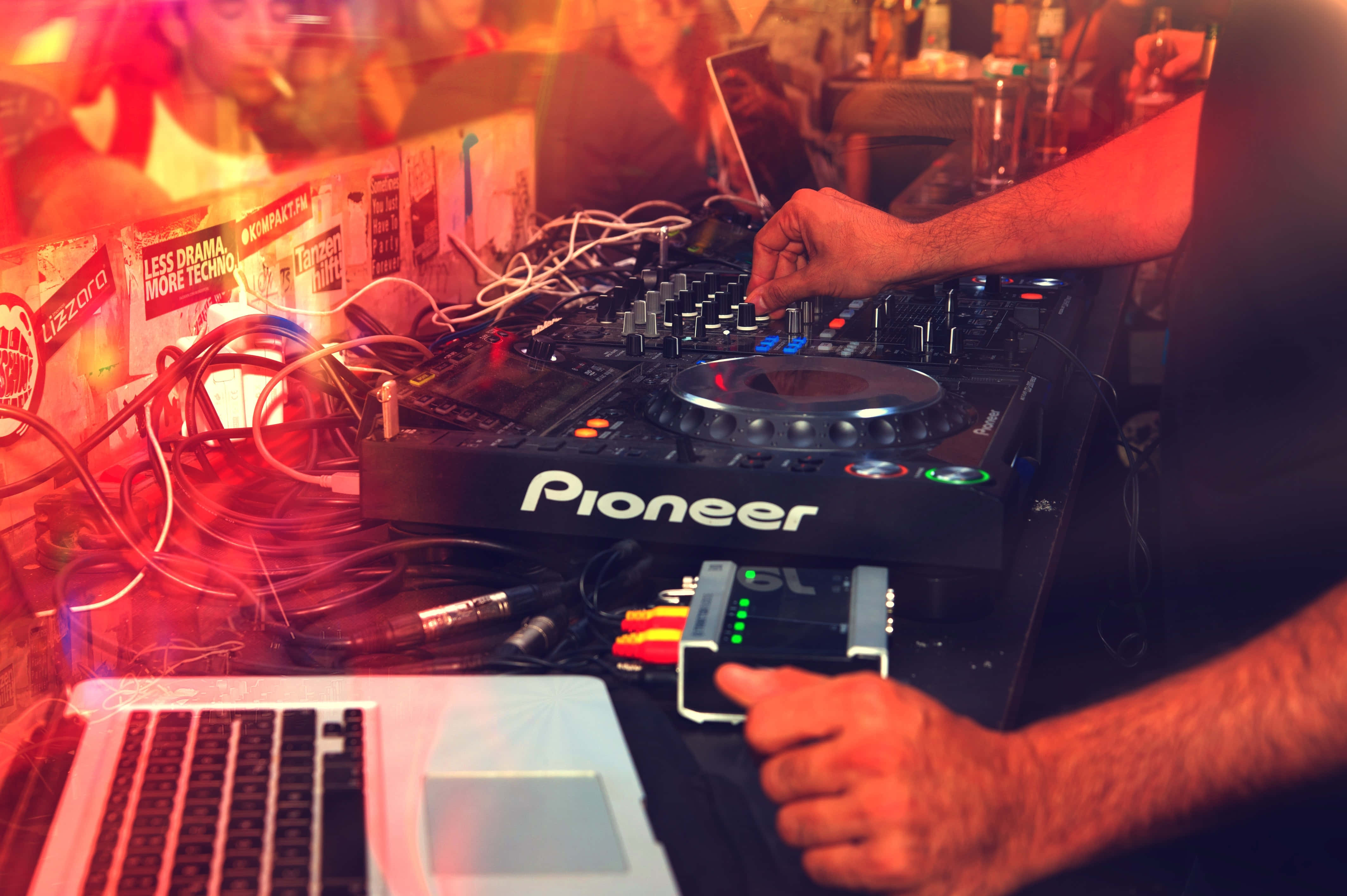 music festival 桌布 壁紙 — 一位DJ的手熟練地操作著Pioneer混音器，在充滿活力的紅色和橙色俱樂部燈光下，旁邊有纏繞的電線和一部手提電腦。