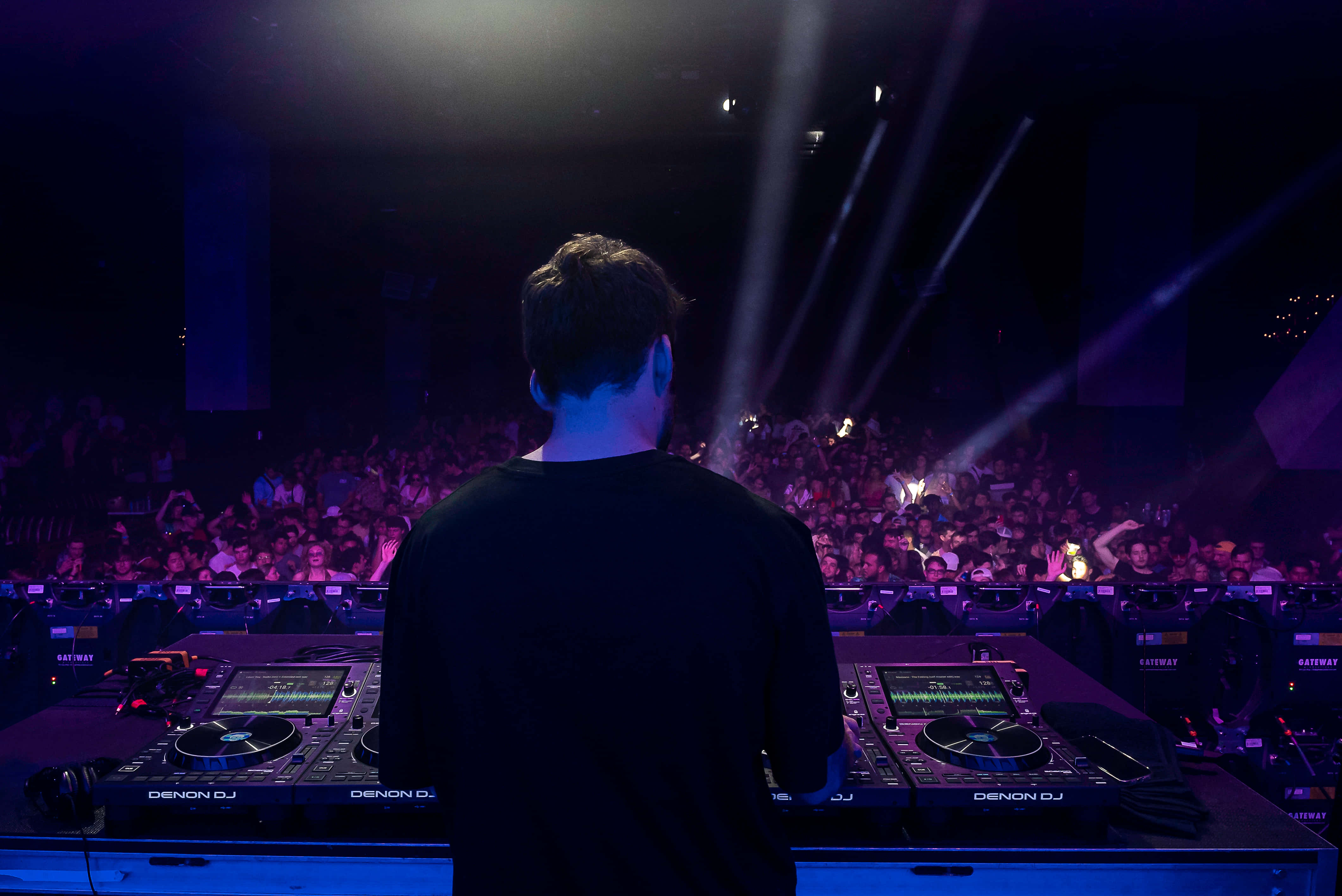 DJ 桌布 壁紙 — 一位DJ站在發光的唱盤設備前，面對擠滿的人群，在戲劇性的紫色舞台燈光下。
