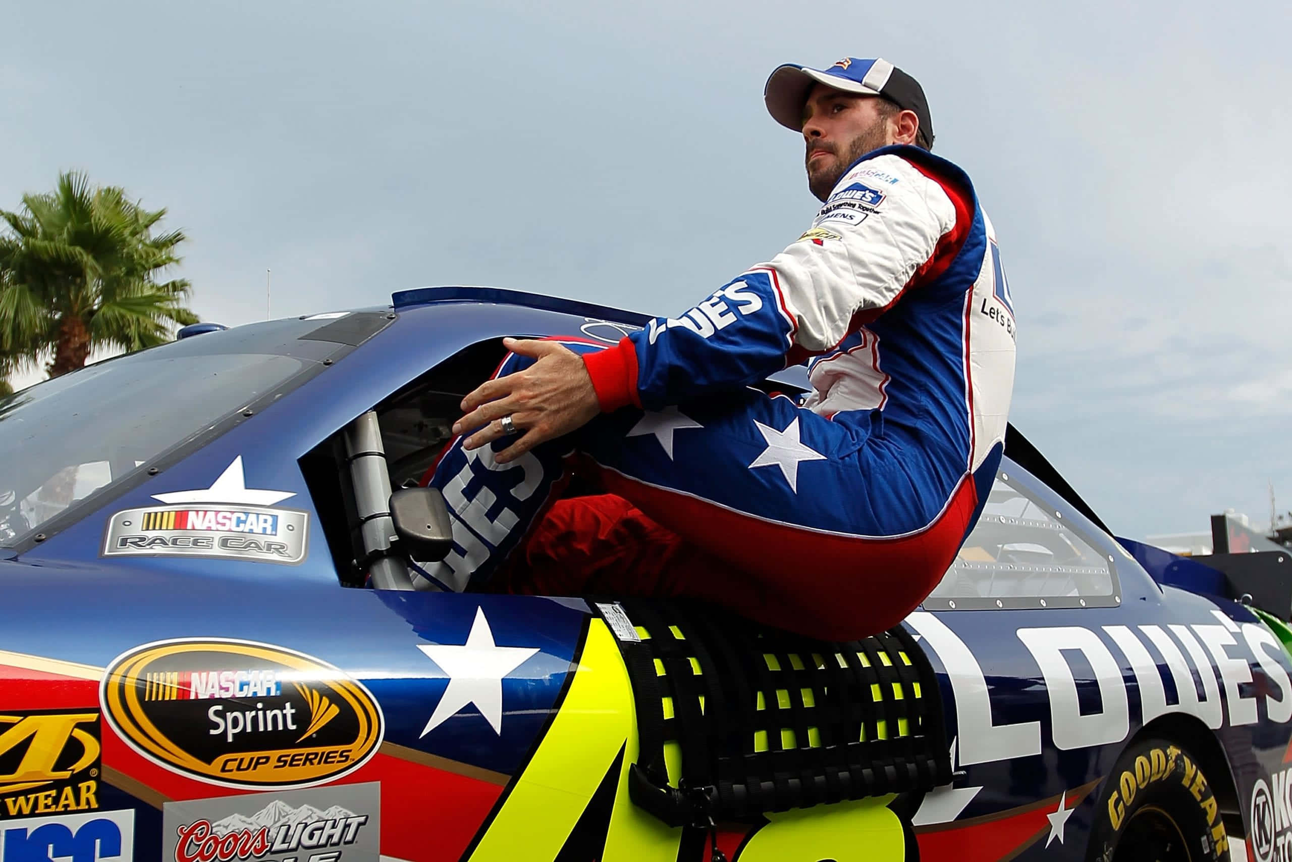 Jimmie Johnson 桌布 壁紙 — 一位NASCAR車手身穿愛國藍紅白賽車服，在多雲的天空下從他裝飾著星星的賽車中探出身子。
