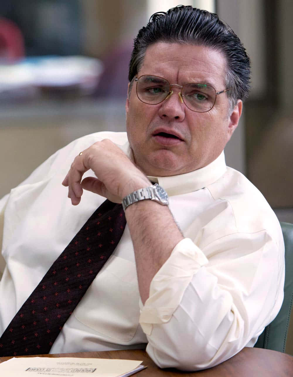Oliver Platt 桌布 壁紙 — 一位中年男士，梳著向後梳的頭髮，戴著眼鏡，坐在辦公桌前，表情驚訝或擔憂。
