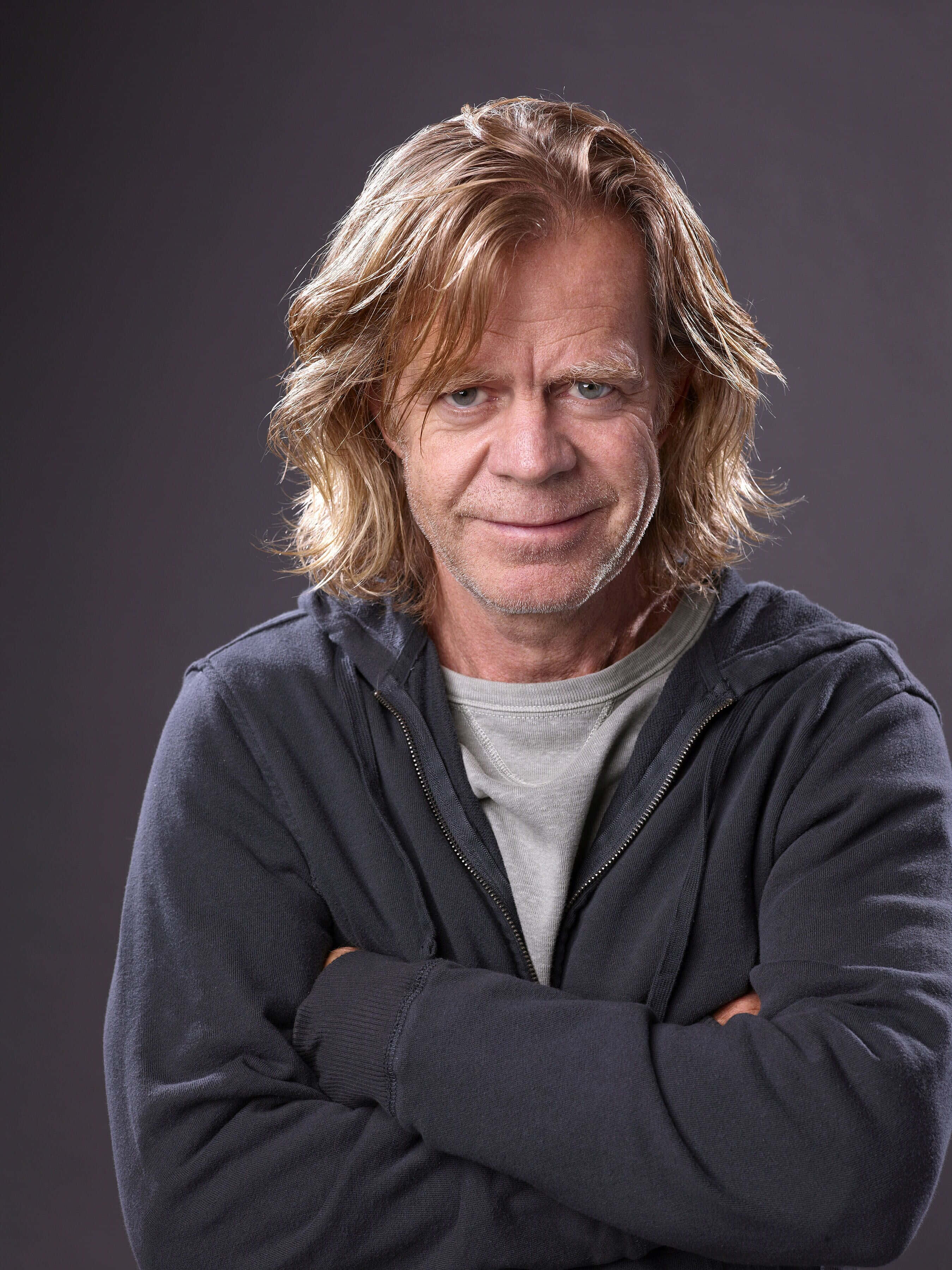 william h. macy 桌布 壁紙 — 一位中年男子，留著及肩的金色長髮，面帶微笑，自信地交叉雙臂站在深灰色工作室背景前。