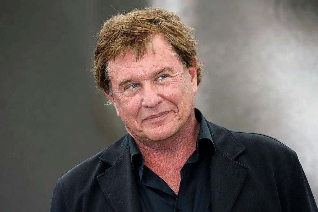Tom Berenger 桌布 壁紙 — 一位中年男子嘅特寫肖像，佢有亂糟糟嘅棕色頭髮同微妙嘅微笑，著住黑色外套，背景係柔和嘅灰色。