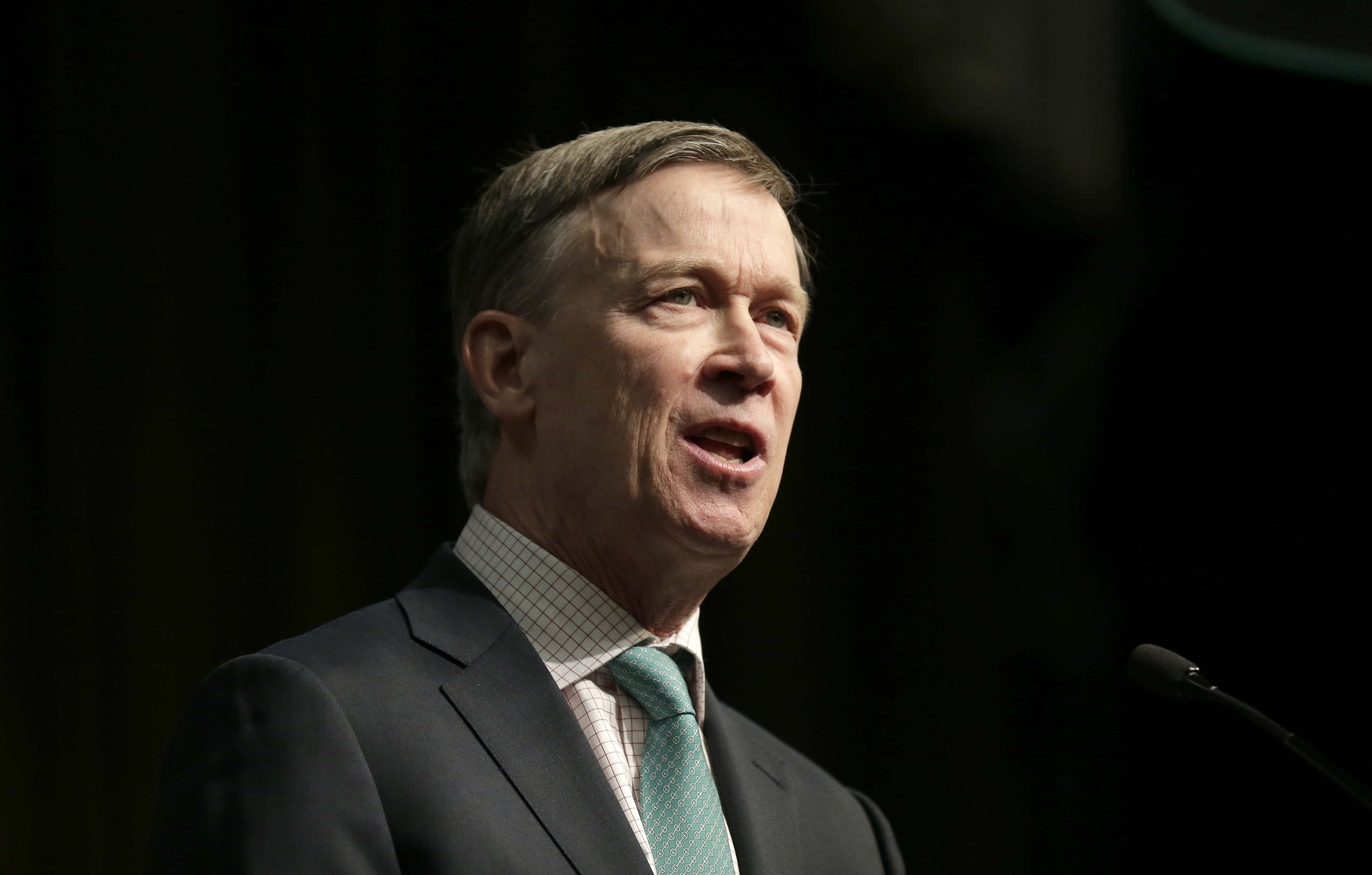 John Hickenlooper Looking Towards Right With Black Background 壁紙 — 一位中年男子穿著深色西裝和綠色領帶，在深色背景下對著麥克風講話。