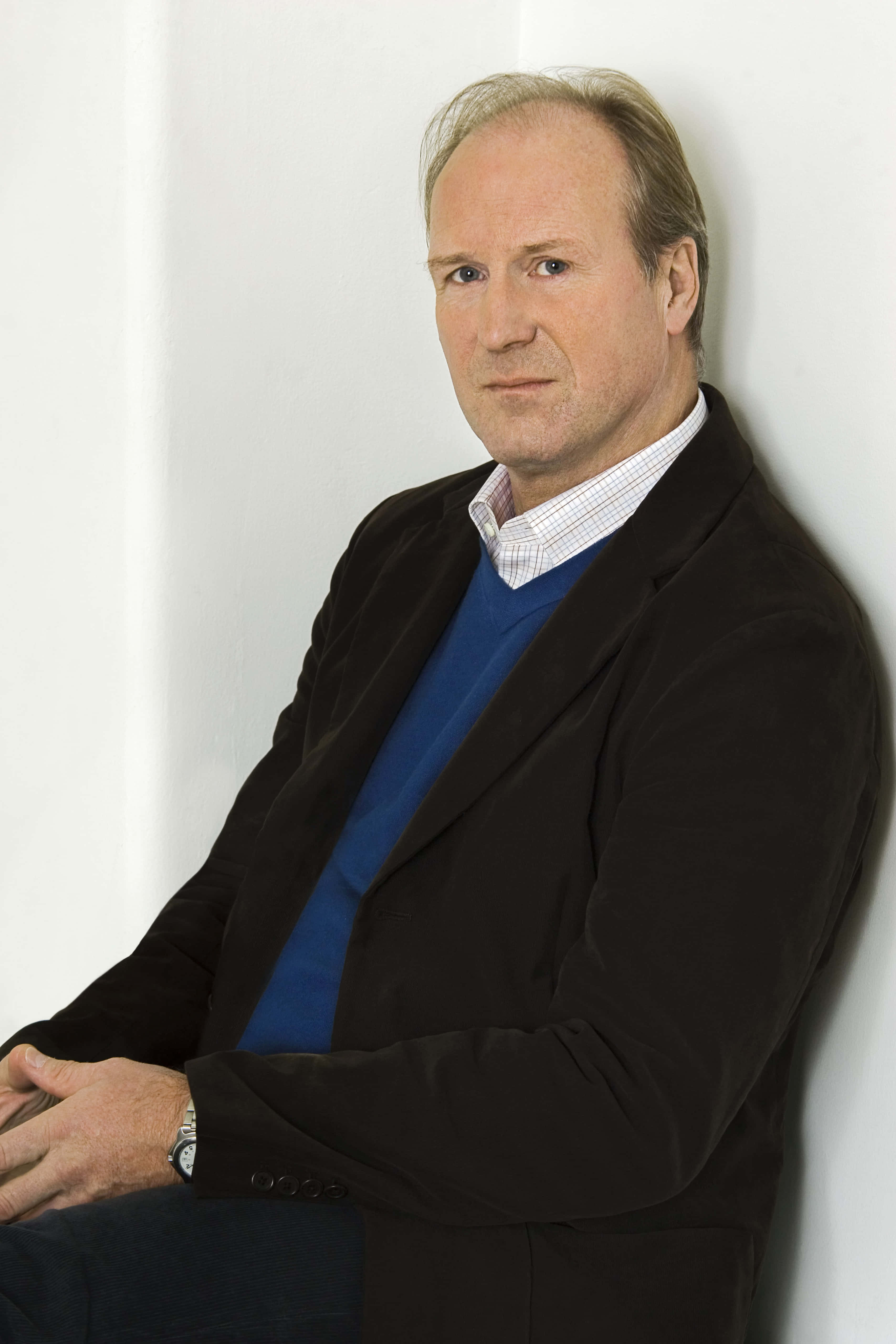 William Hurt 桌布 壁紙 — 一位中年男子身穿深色西裝外套和藍色毛衣，神情平靜地靠在一面純白色的牆上。