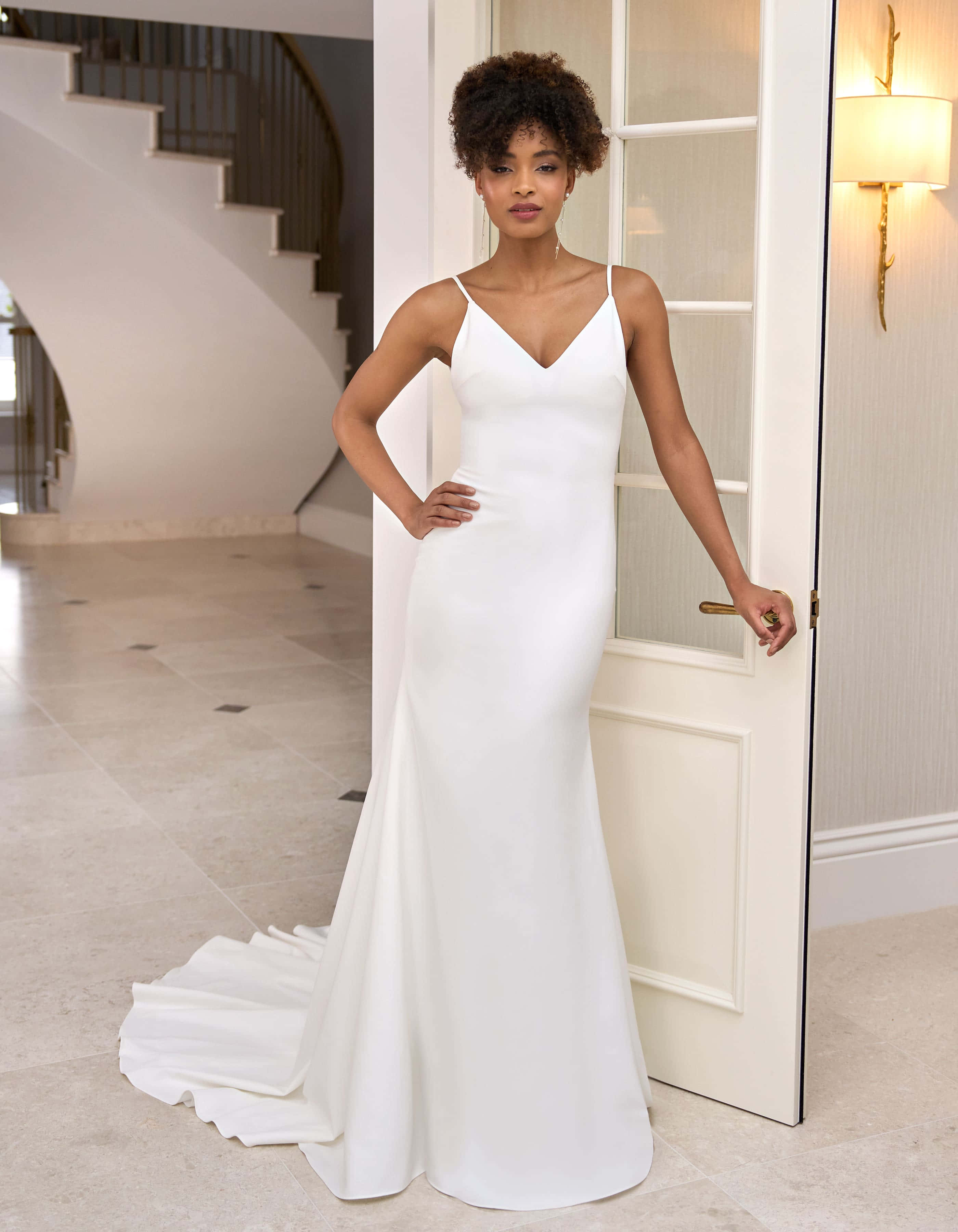 Bride's Dress 桌布 壁紙 — 一位優雅嘅女人著住一件修身白色婚紗，喺一間有弧形樓梯嘅現代住宅入面，優雅咁企喺一道開咗嘅門旁邊。
