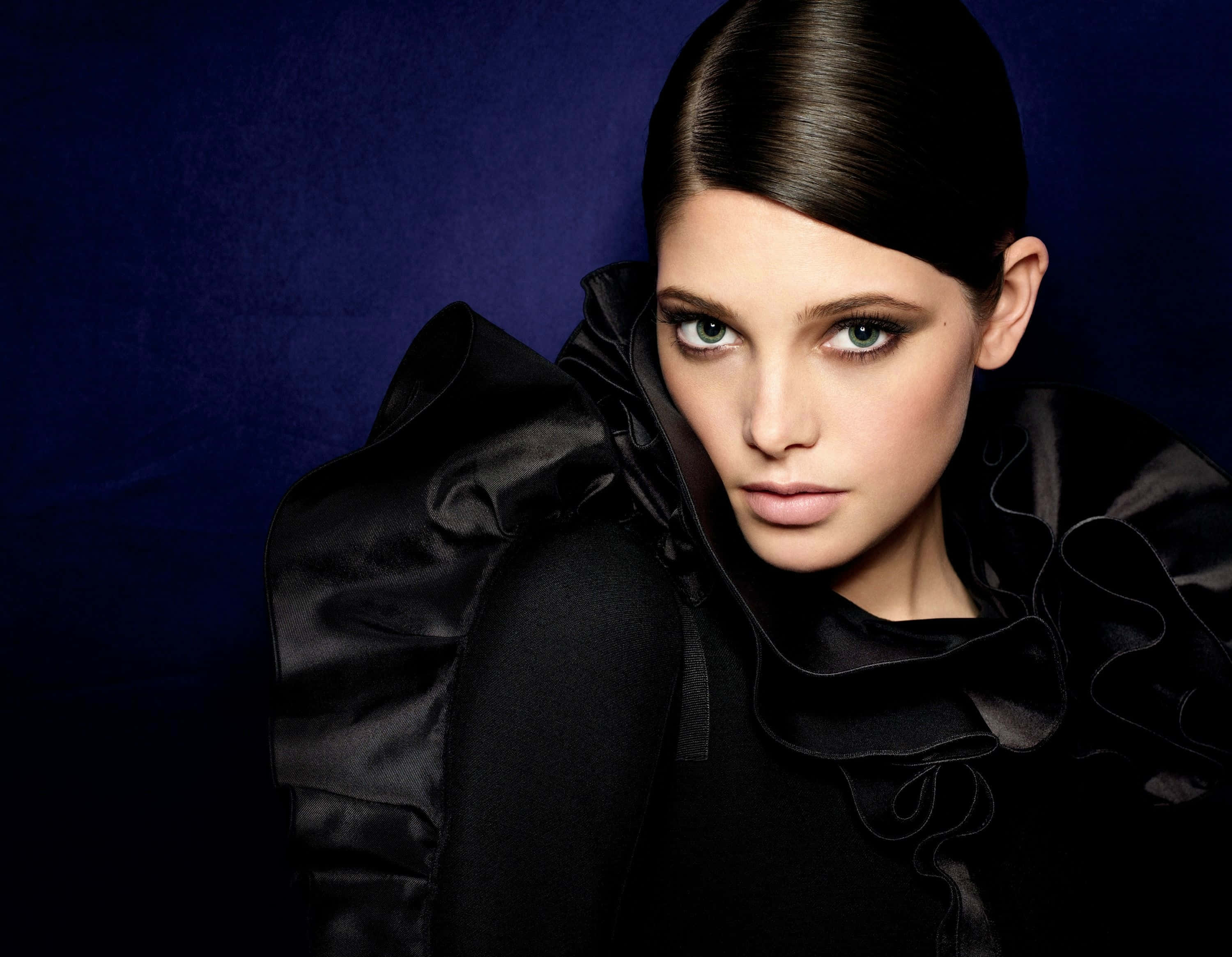 ashley greene 桌布 壁紙 — 一位優雅的女人，擁有引人注目的綠色眼睛和光滑的黑髮，穿著一件戲劇性的黑色荷葉邊服裝，背景是深藍色紋理背景。