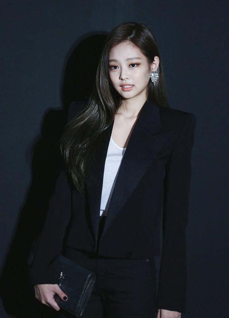 Jennie Kim 桌布 壁紙 — 一位優雅的女士身穿利落的黑色西裝外套和白色上衣，站在深色背景前，手持手拿包，佩戴閃亮耳環。