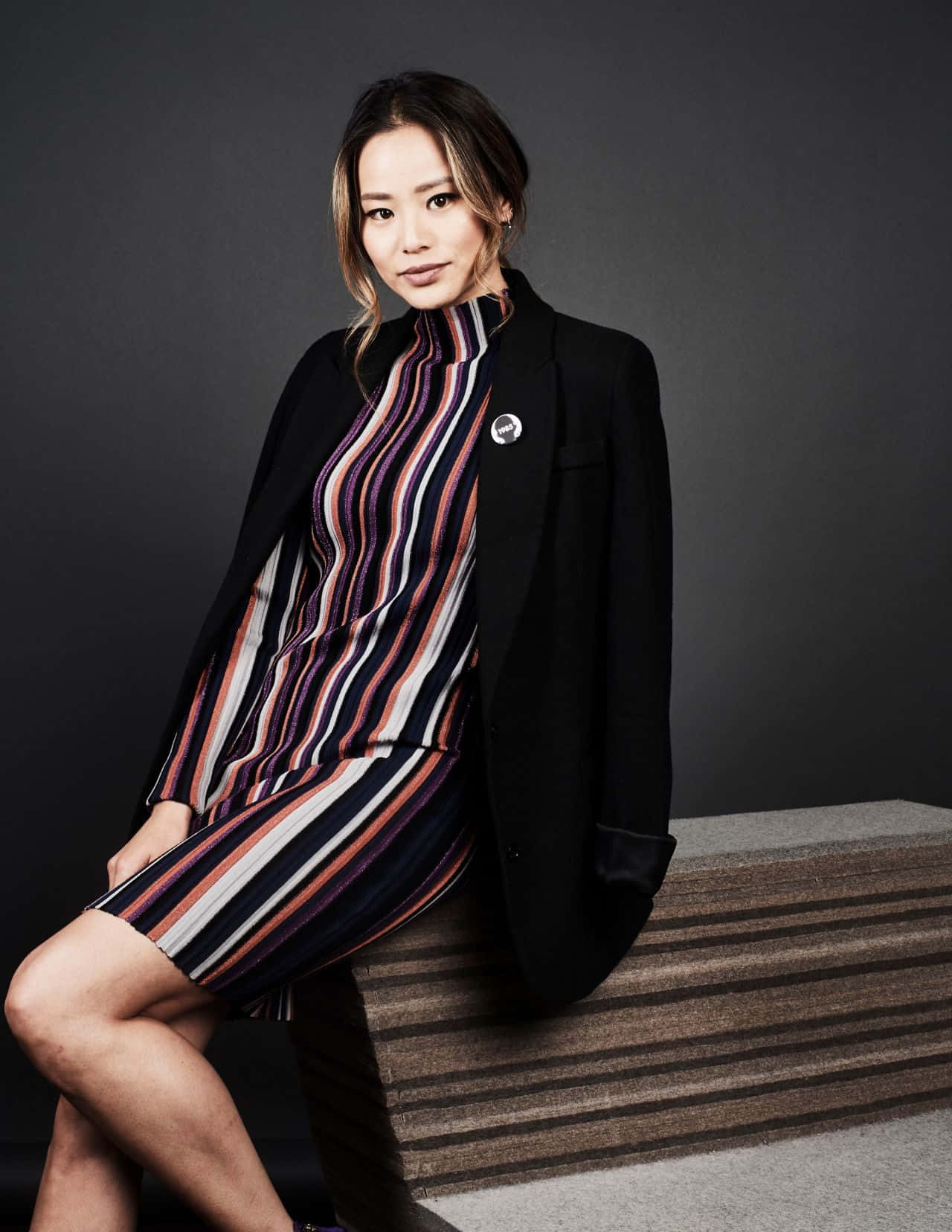 Jamie Chung 桌布 壁紙 — 一位優雅的女士身穿黑色西裝外套和條紋連衣裙，自信地坐在質感長椅上，背景是深色攝影棚。