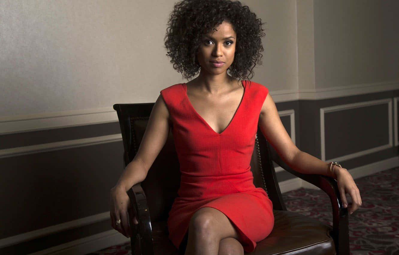Gugu Mbatha-raw 桌布 壁紙 — 一位優雅的捲髮女性坐在深色椅子上，身穿搶眼的紅色連衣裙，背景是柔和的牆壁。