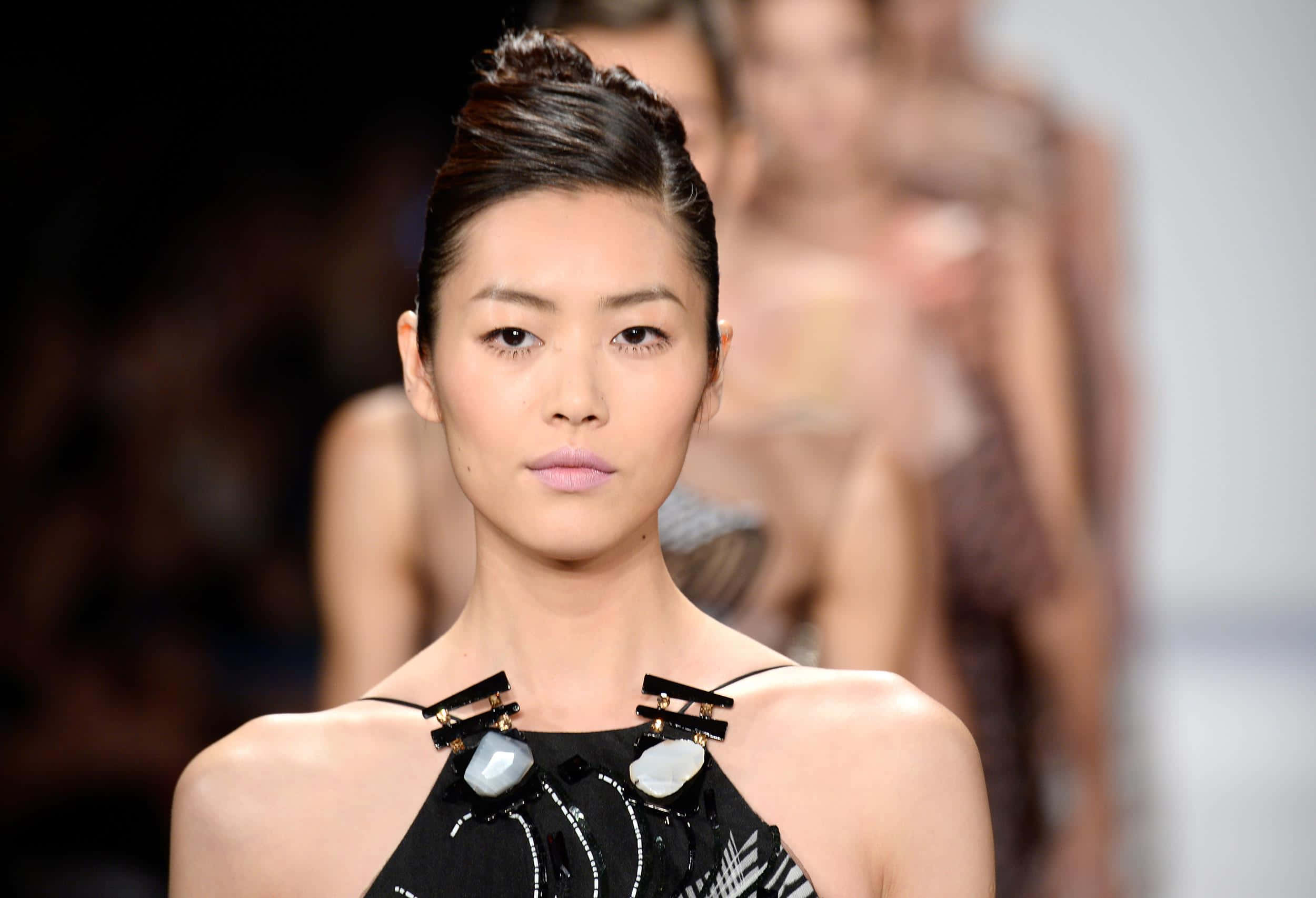 Beautiful Fashion 桌布 壁紙 — 一位優雅的模特兒梳著光滑的髮髻，佩戴大膽的珠寶，在明亮的燈光下自信地走在時裝天橋上。