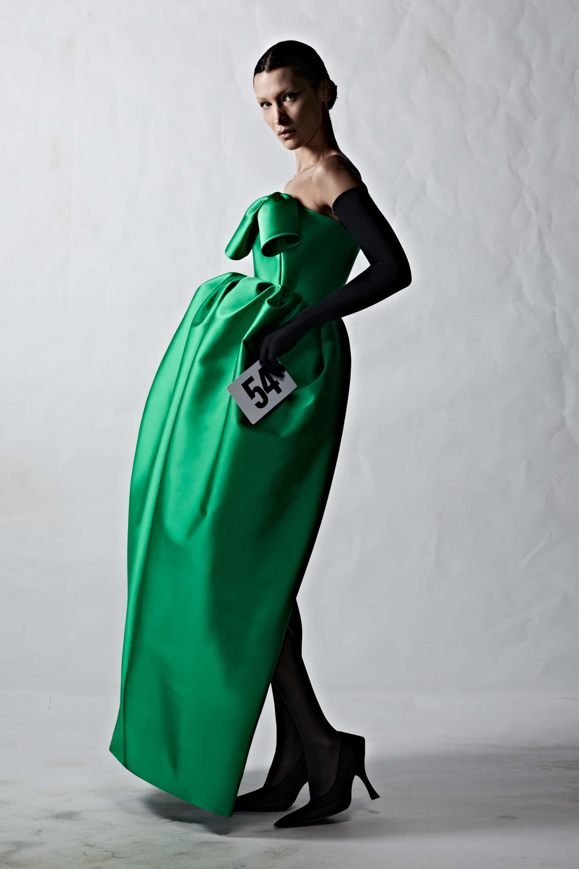 Green Fashion 桌布 壁紙 — 一位優雅的模特兒穿著搶眼的翡翠綠緞面禮服，配有大蝴蝶結和黑色配飾，在帶有紋理的白色背景前擺姿勢。