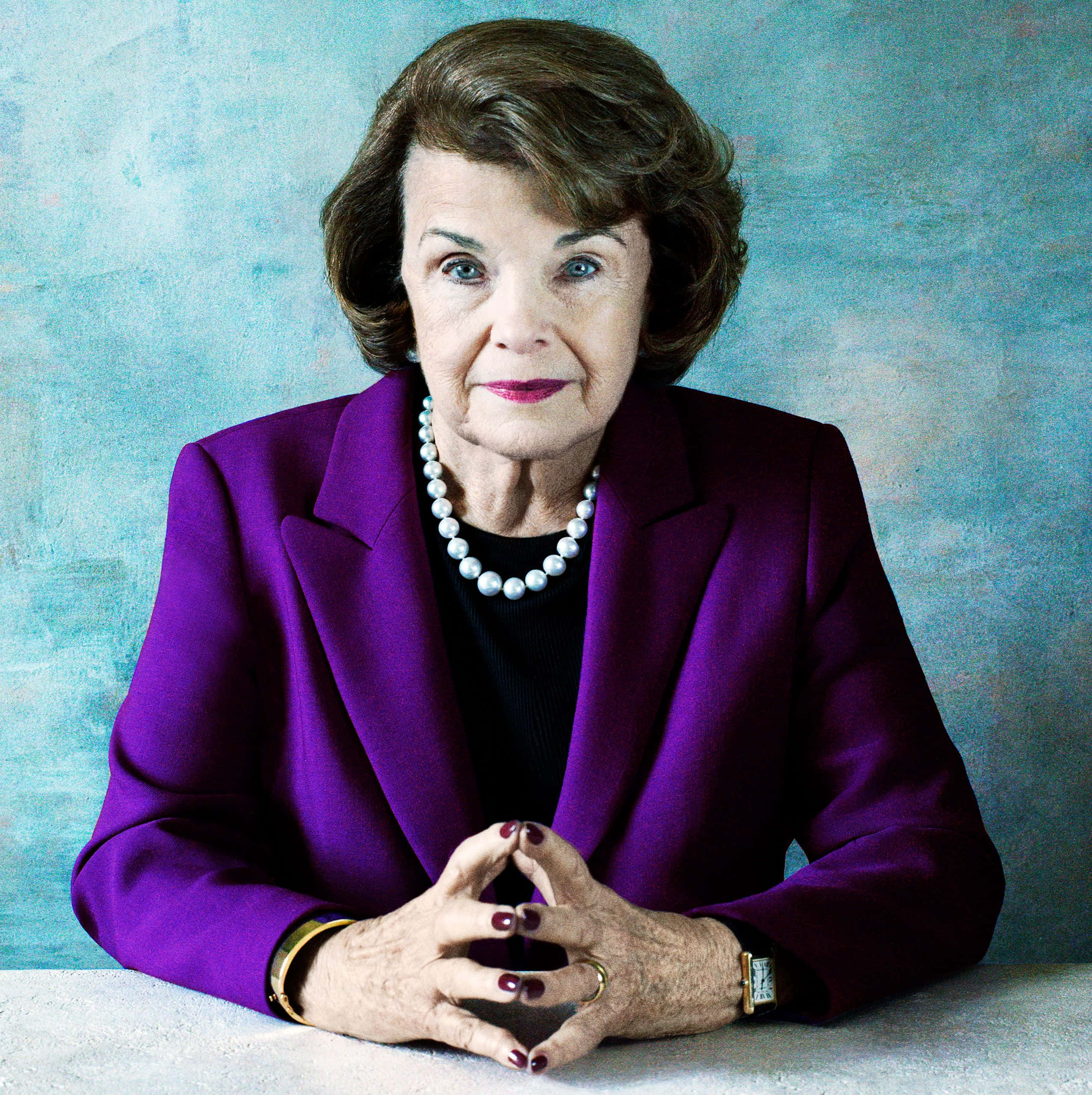 Dianne Feinstein 桌布 壁紙 — 一位優雅的老年女士身穿鮮豔紫色西裝外套和珍珠項鍊，雙手交疊在桌上，直視鏡頭，背景為有紋理的藍色。