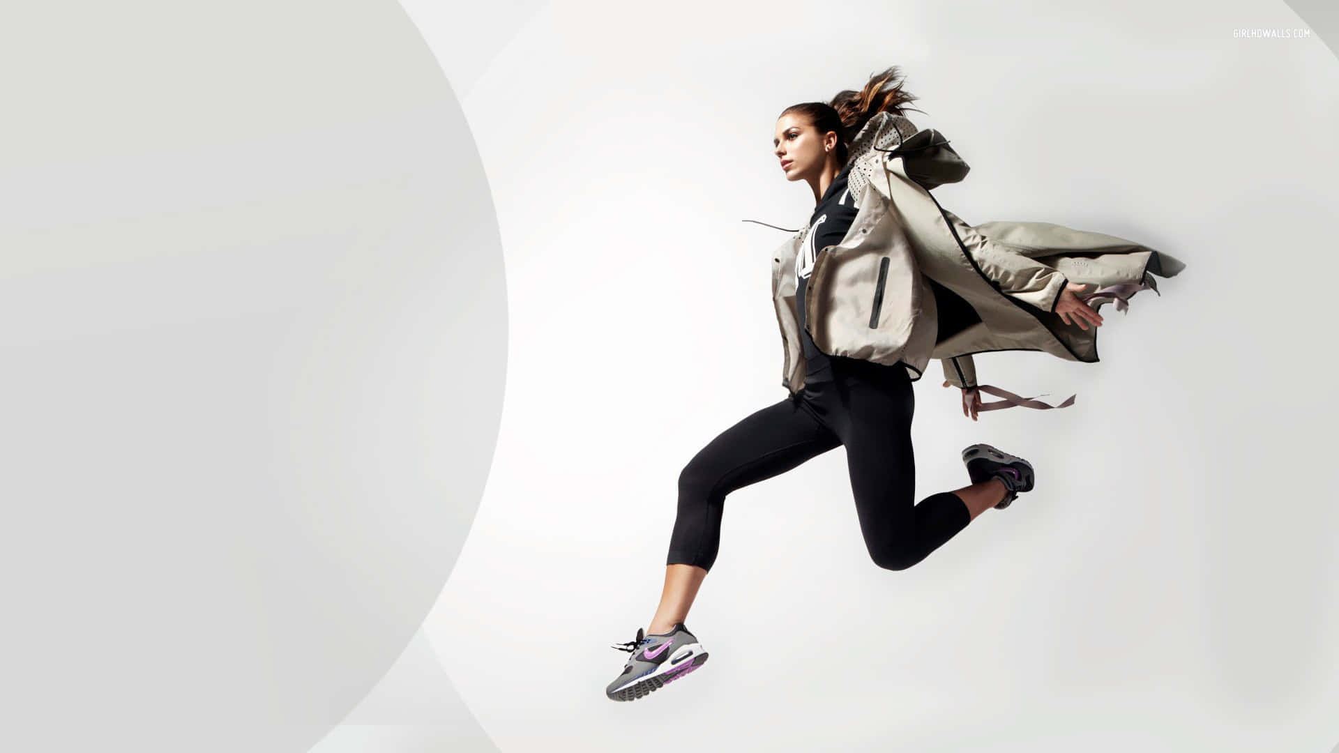 Alex Morgan 桌布 壁紙 — 一位充滿活力的女性穿著運動裝備，在簡約的白色背景前騰空跳躍，外套飄逸。