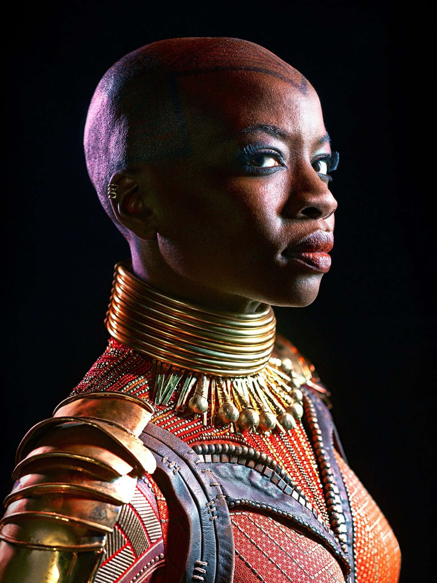 Okoye 桌布 壁紙 — 一位兇猛的女戰士女王，身穿華麗的金色盔甲和紅色珠飾服裝，在黑暗背景下專注地凝視。
