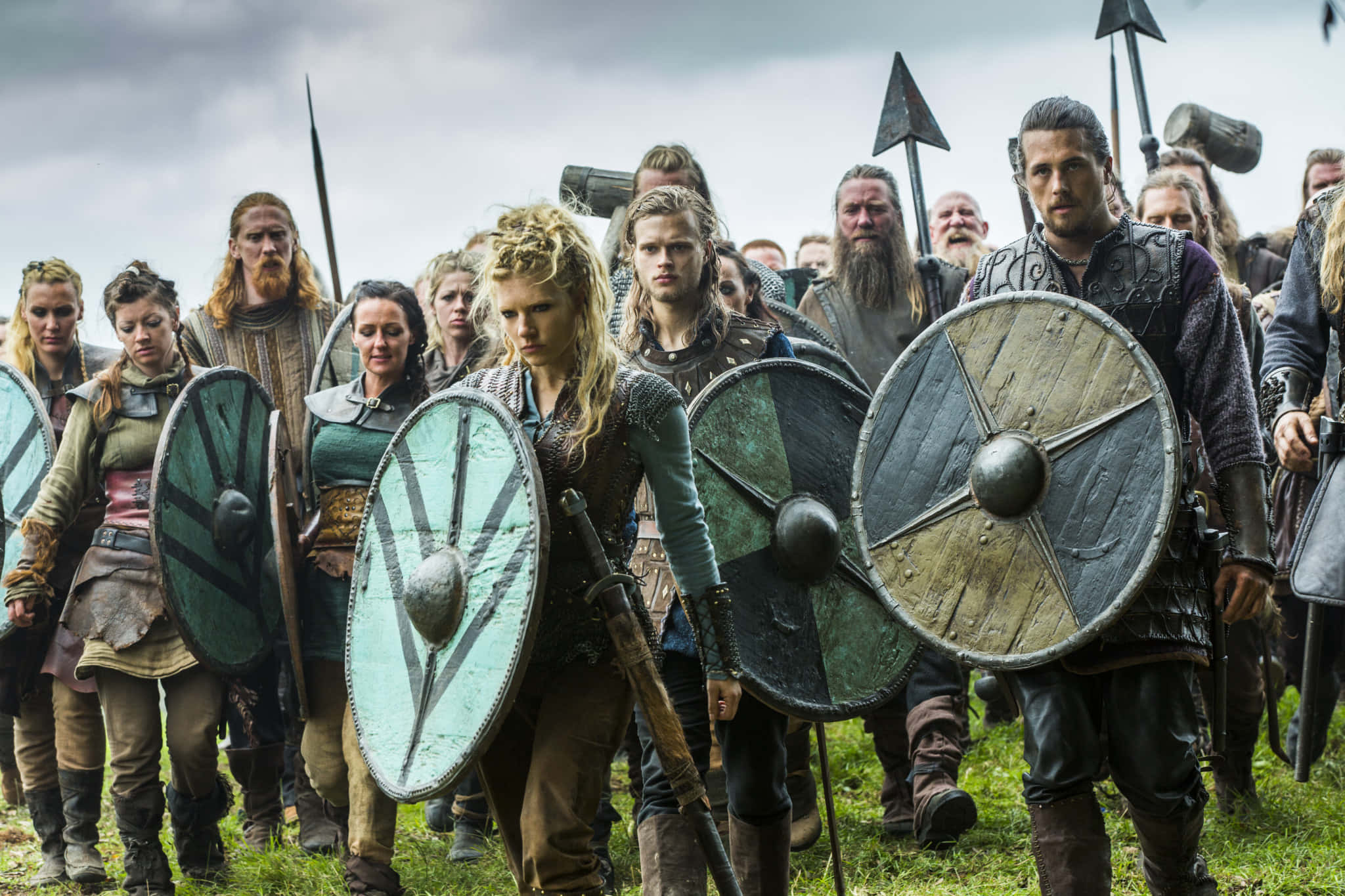 Vikings 桌布 壁紙 — 一位兇猛的維京女戰士帶領著一個準備戰鬥的部族，手持圓盾和長矛，在陰雲密佈的天空下。
