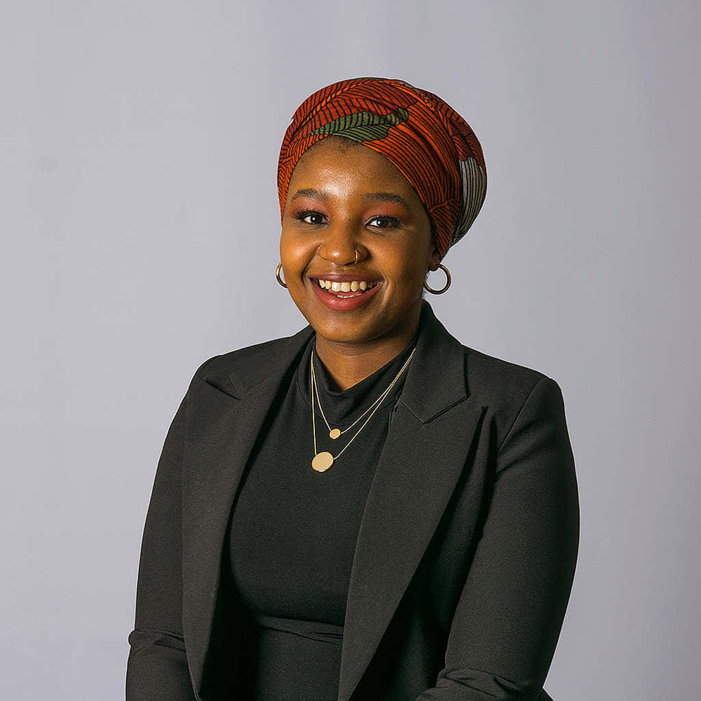 African 桌布 壁紙 — 一位光彩照人的女士，戴著充滿活力的橙色圖案頭巾，身穿時尚黑色西裝外套，在柔和的灰色背景下溫暖微笑。
