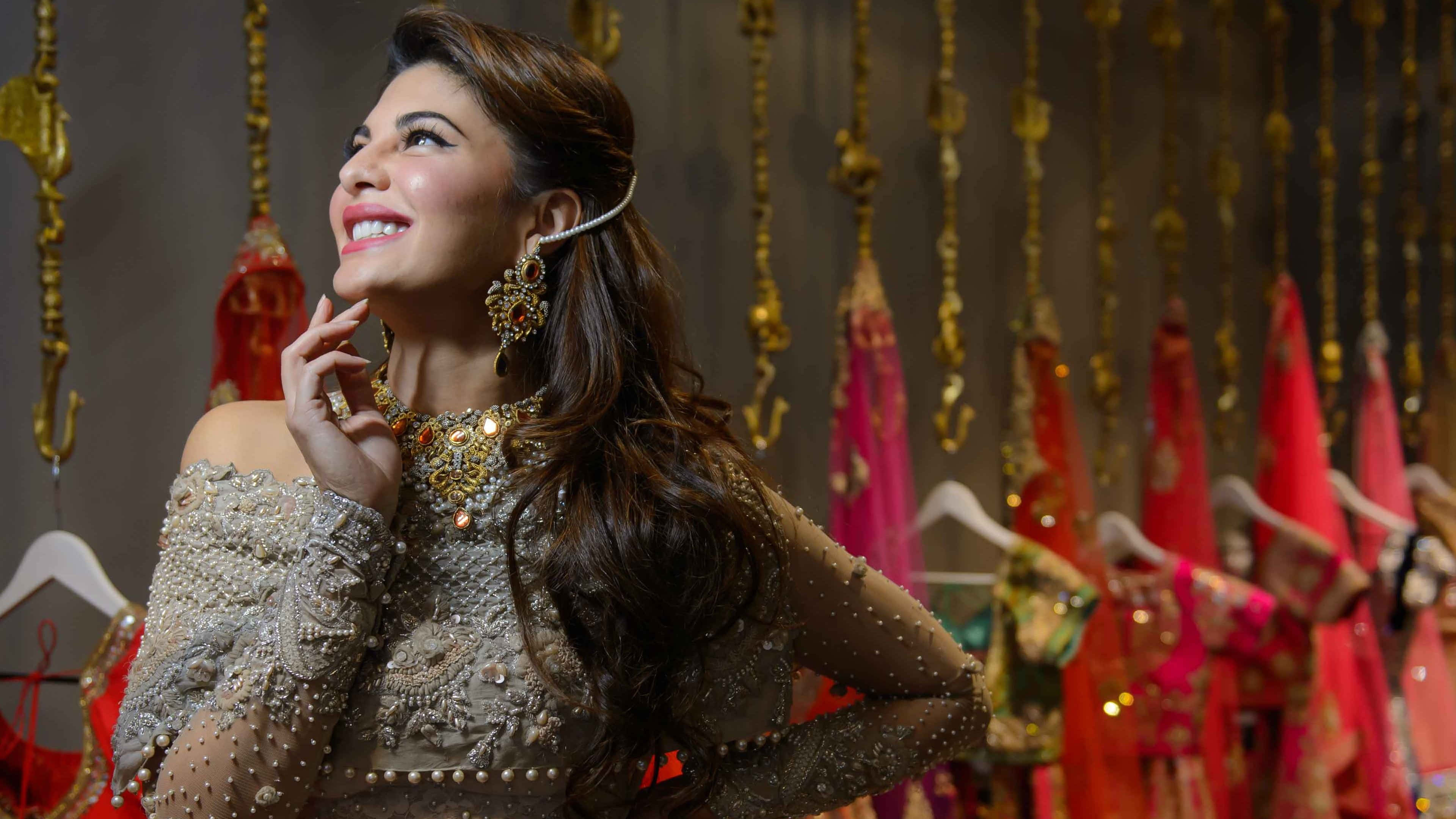 Jacqueline Fernandez 桌布 壁紙 — 一位光彩照人的女性，佩戴華麗的金飾和裝飾服飾，在懸掛的傳統服裝中向上微笑。